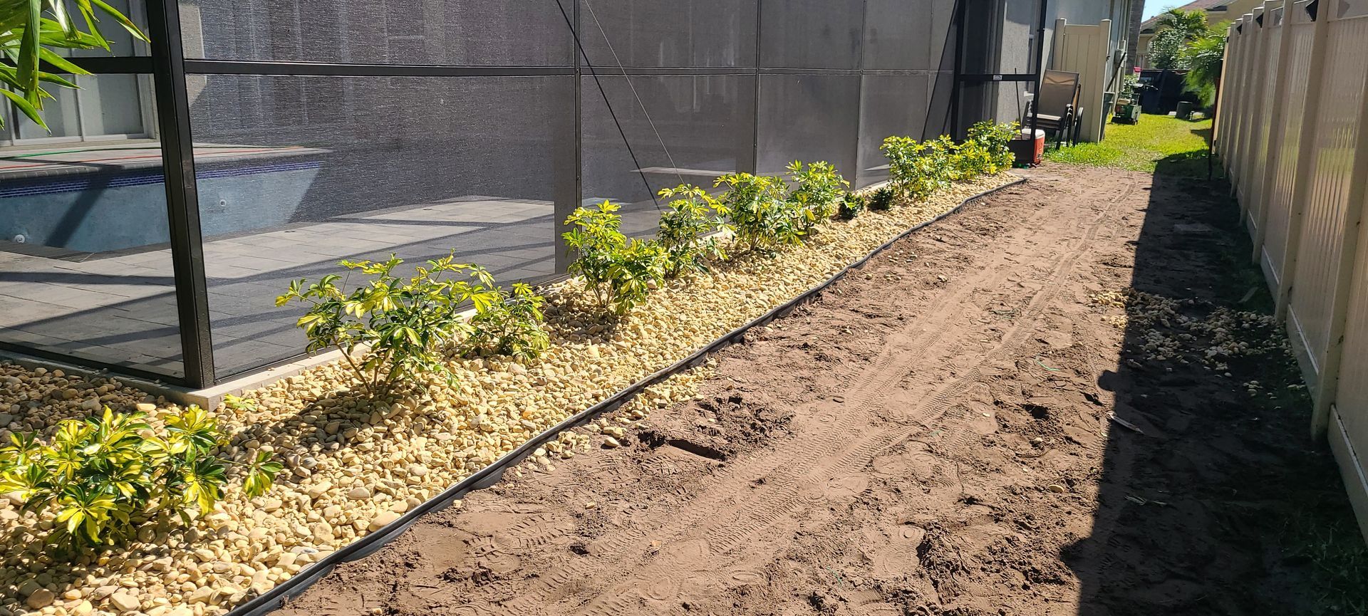 A landscaped garden bed with small green shrubs and beige gravel beside a path, adjacent to a mesh screen and fence.