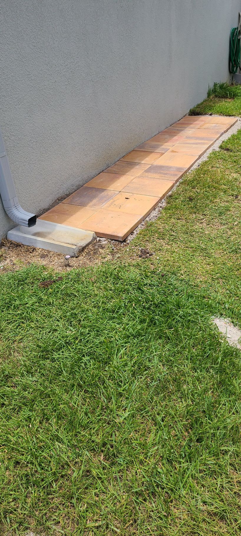 A strip of orange stone pavers along a stucco wall, with a downspout draining onto a concrete block on a grassy lawn.