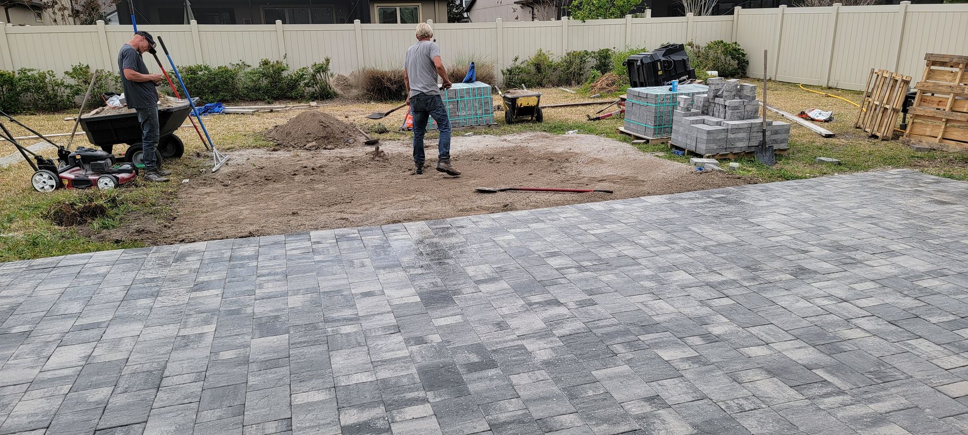 Two workers preparing a backyard area with pavers, tools, and construction materials in a residential setting.