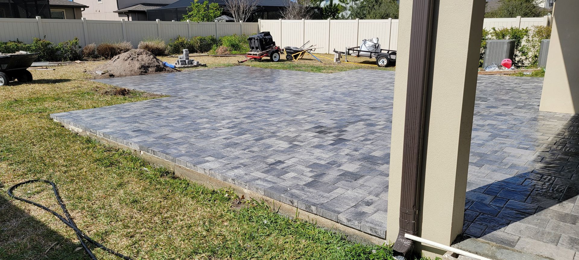 A newly installed grey paver patio in a backyard, viewed from near a tan structural pillar.
