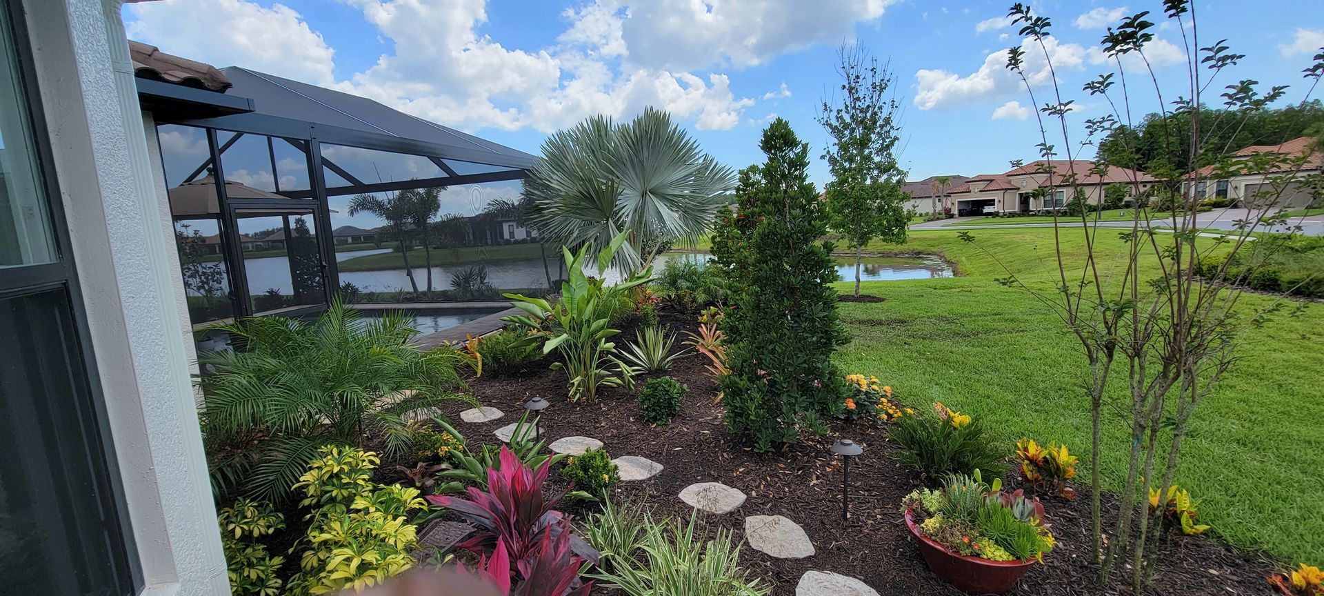 A landscaped garden bed with colorful shrubs and stepping stones, overlooking a screened-in pool deck and pond.