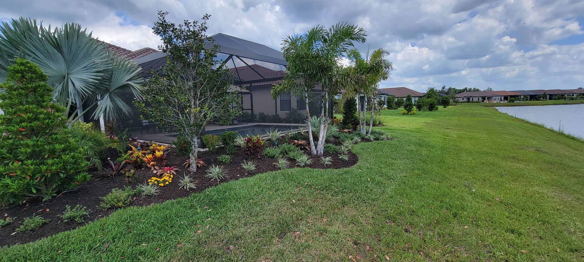 A landscaped garden bed with tropical plants and palm trees sits in front of a screened patio beside a lake.