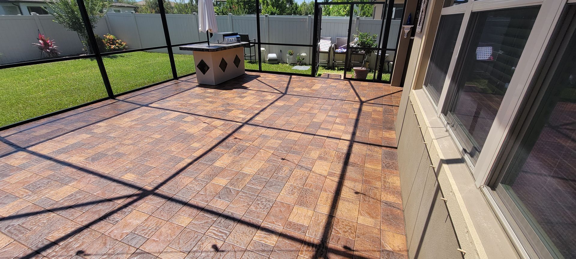 A screened-in patio with patterned brown pavers, looking out toward a grassy yard and a white fence.