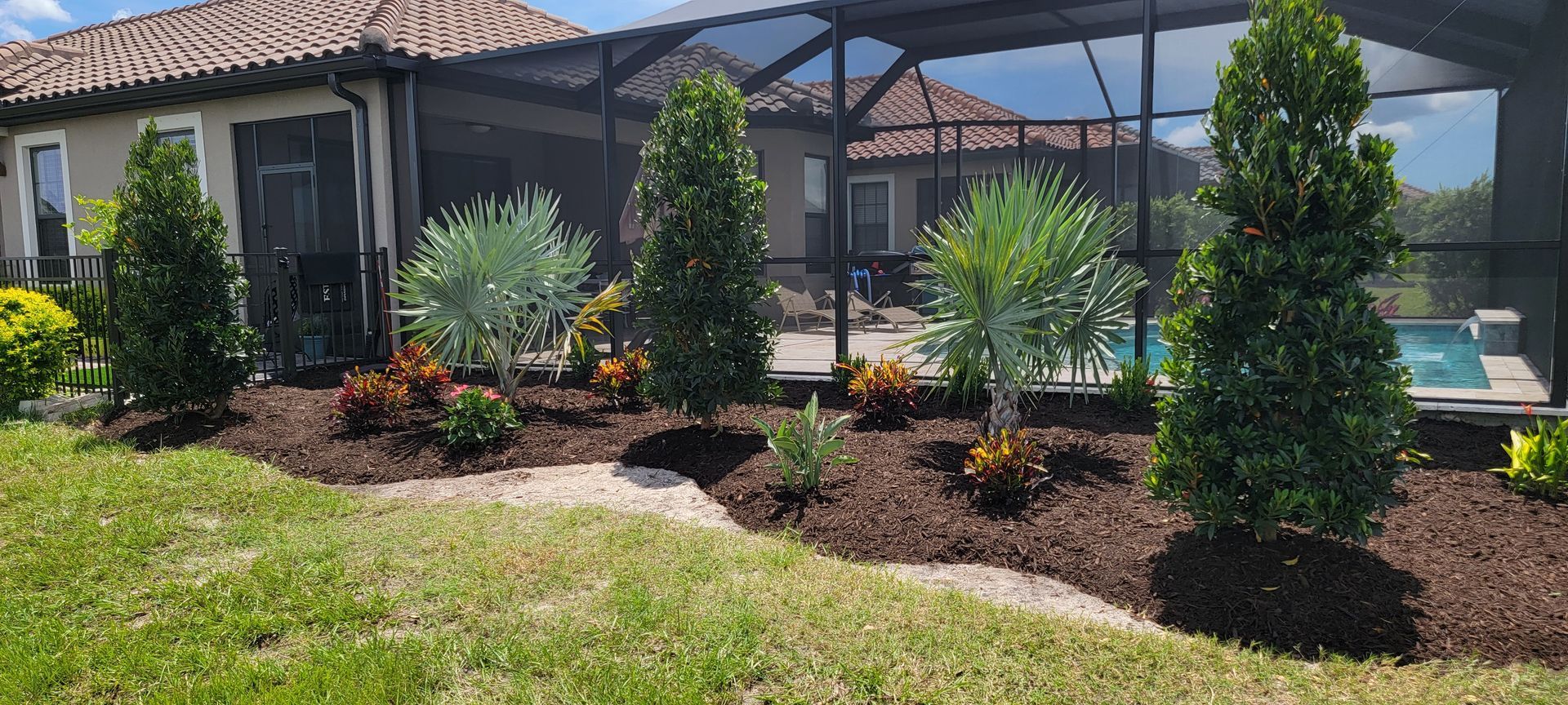 A landscaped garden bed with mulch, shrubs, and small trees fronting a house with a screened-in pool enclosure.