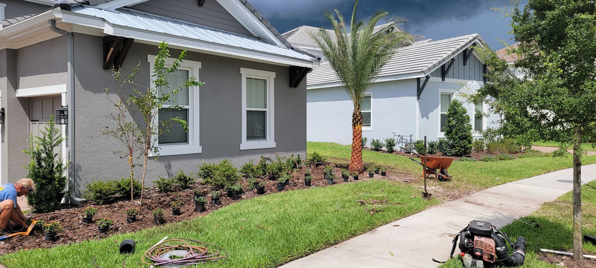 A landscaper plants small shrubs in mulch beds outside a gray residential home on a sunny day.