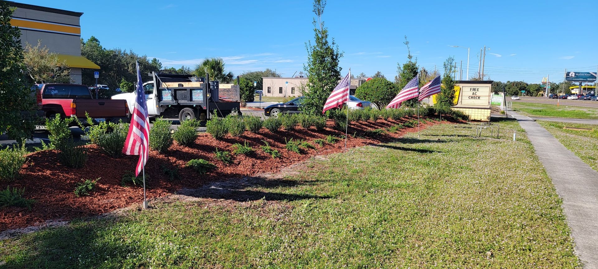 Small American flags line a landscaped garden bed with mulch and shrubs in front of a parking lot on a sunny day.