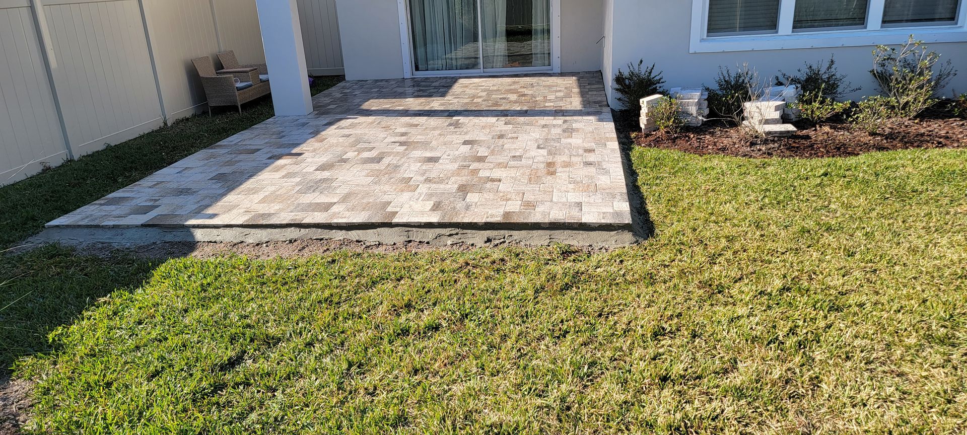 A tan and gray paved backyard patio extending from a house, featuring a white pillar and adjacent landscaped garden bed.