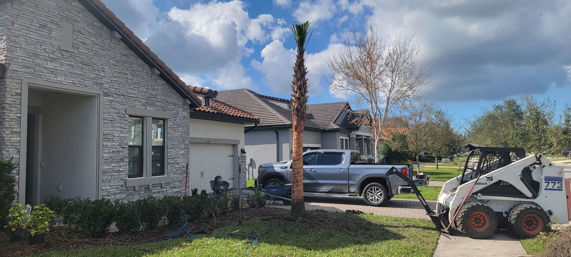A white skid steer loader parked on a sidewalk in front of a residential house with a single palm tree in the front yard.