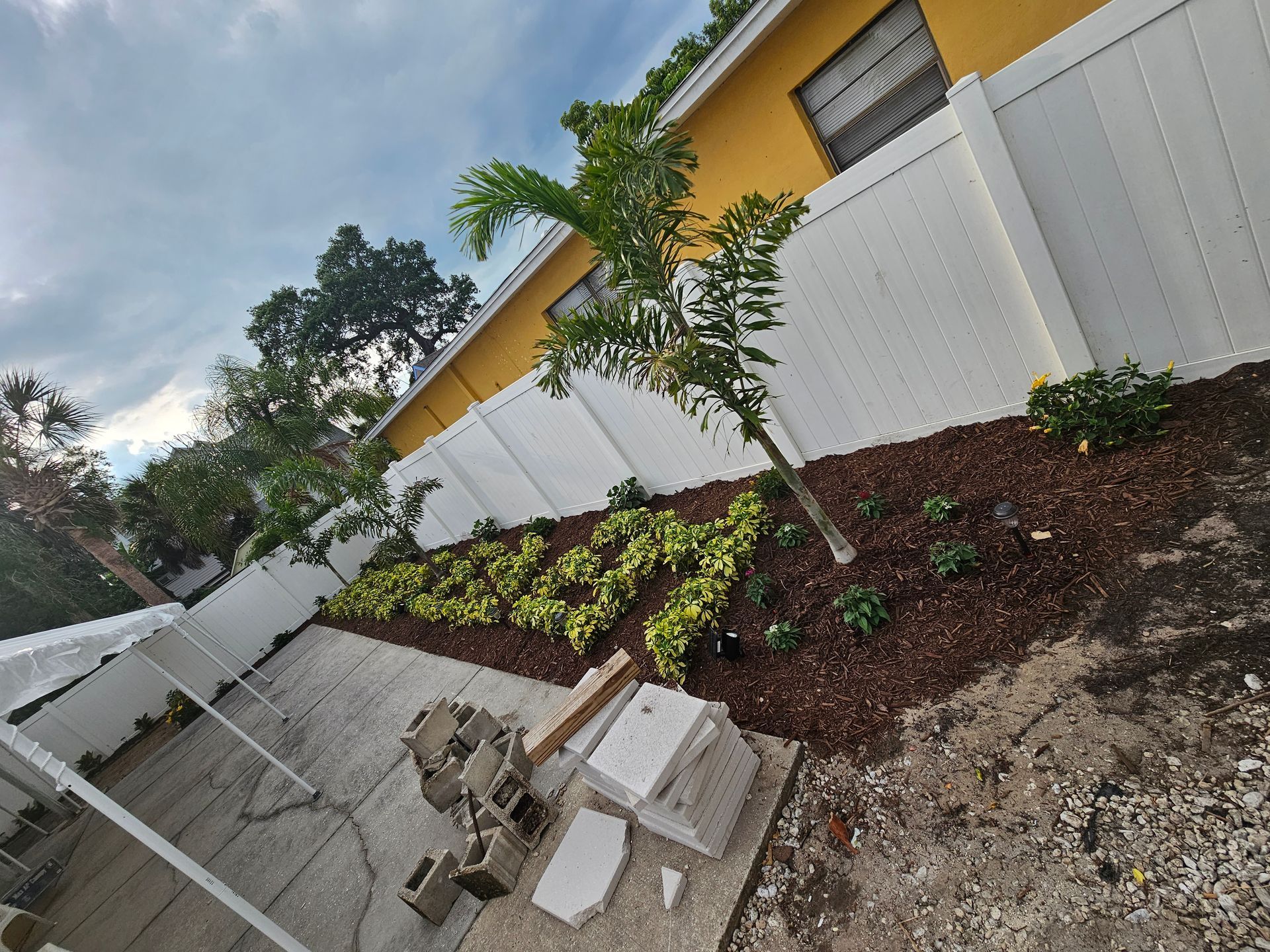 A backyard scene featuring a white vinyl fence, a small palm tree, mulched garden beds with plants, and stacked pavers.