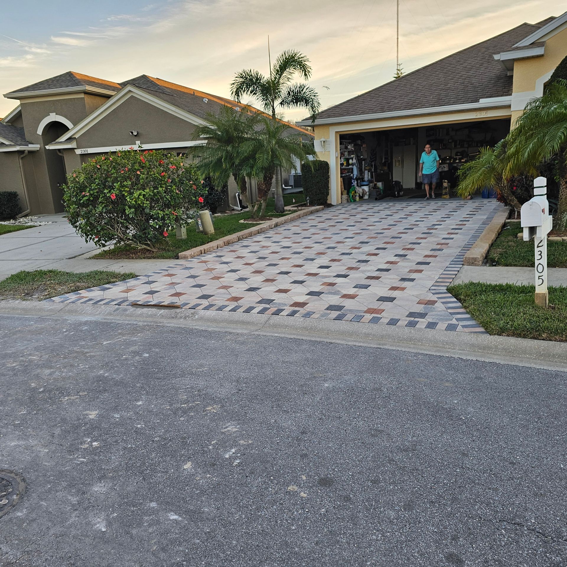 A paved driveway made of multi-colored square tiles sits in front of a house, with a person standing inside the garage.