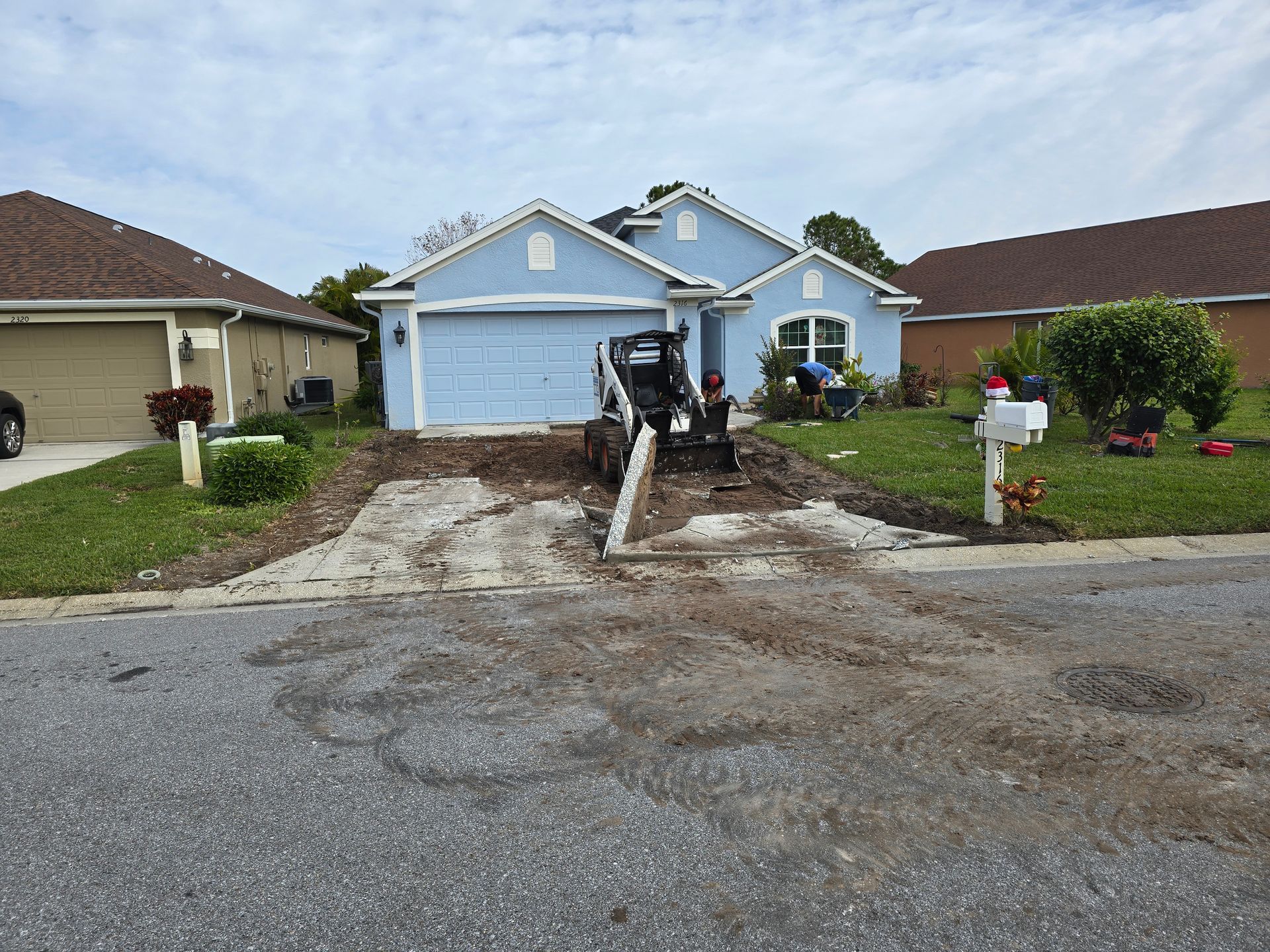 A skid steer sits on a torn-up driveway in front of a blue single-story house during construction.
