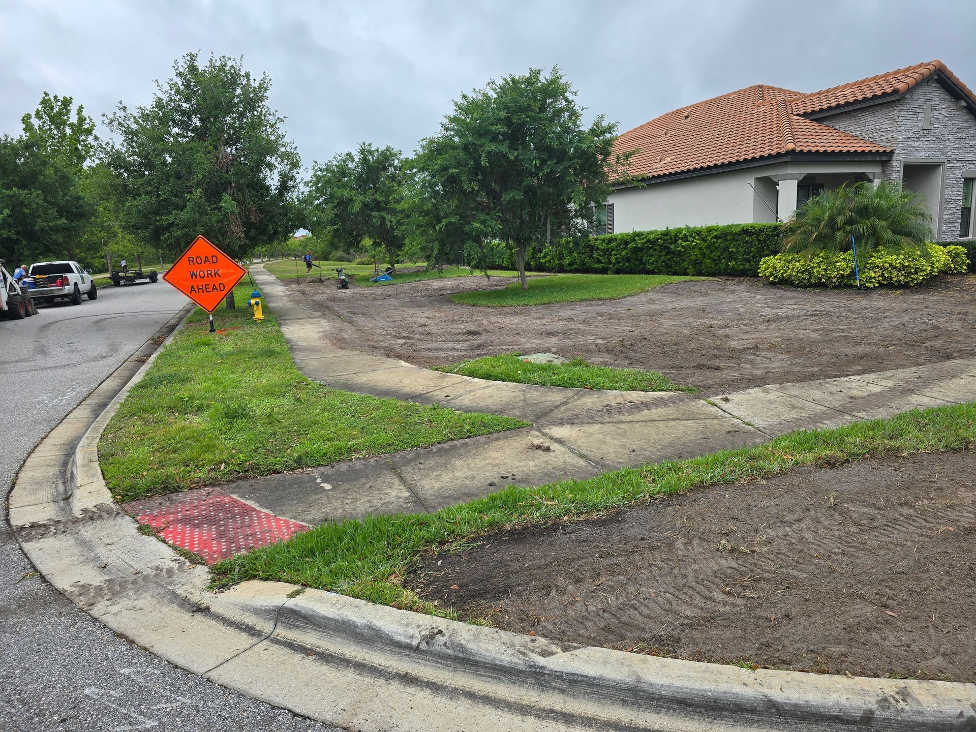 An orange road work sign sits near a curb on a residential street next to a house with an empty, recently cleared yard.