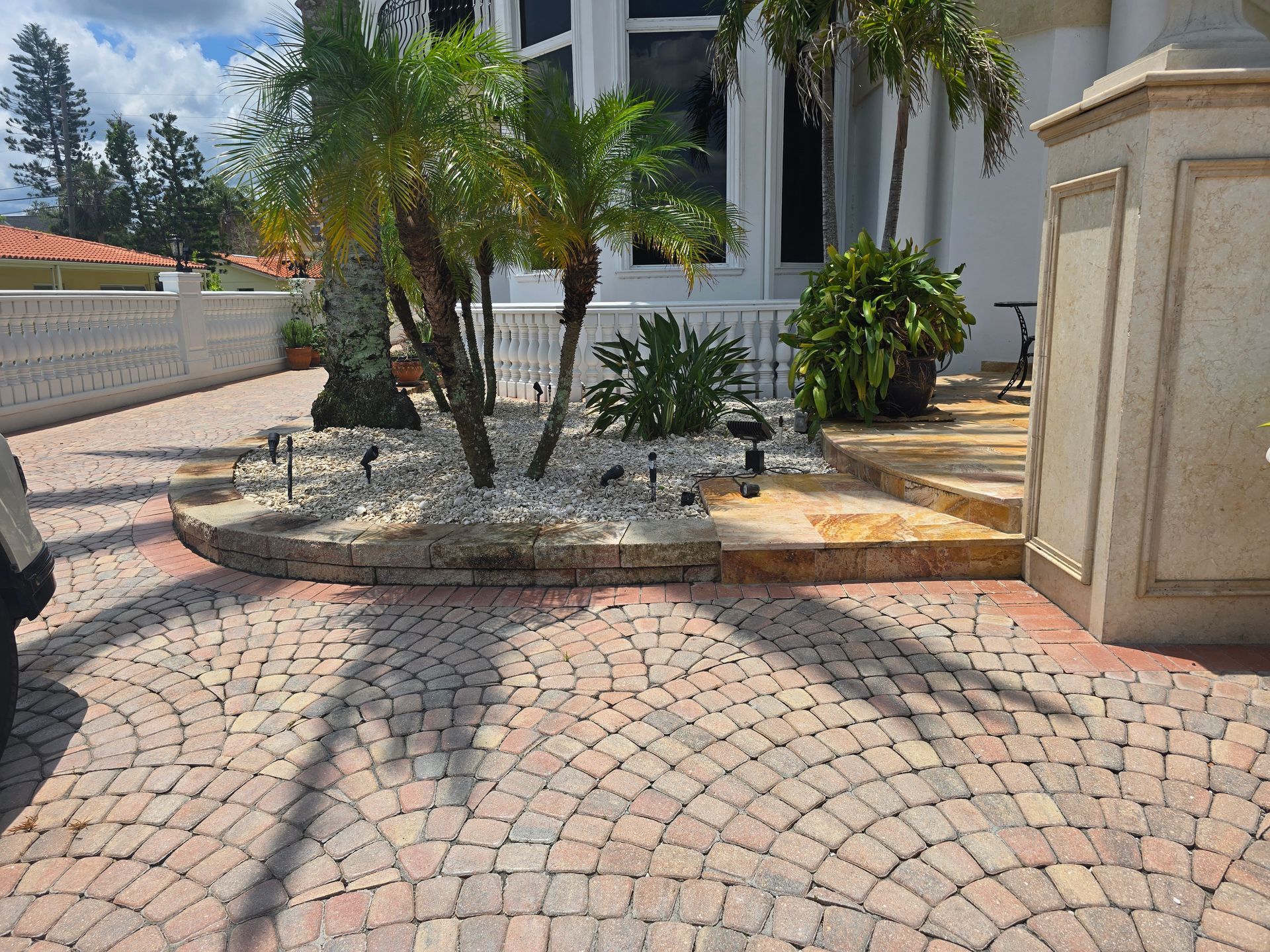 A paved circular driveway leading to a landscaping bed with small palm trees and lush greenery in front of a white house.