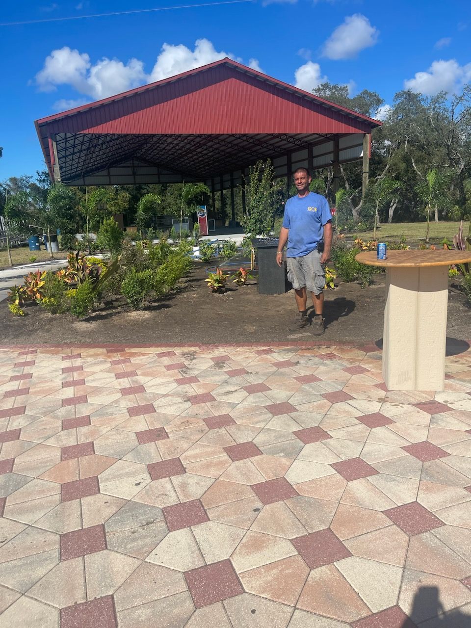 A person stands in a landscaped outdoor area with a covered pavilion in the background and a pedestal table to the right.