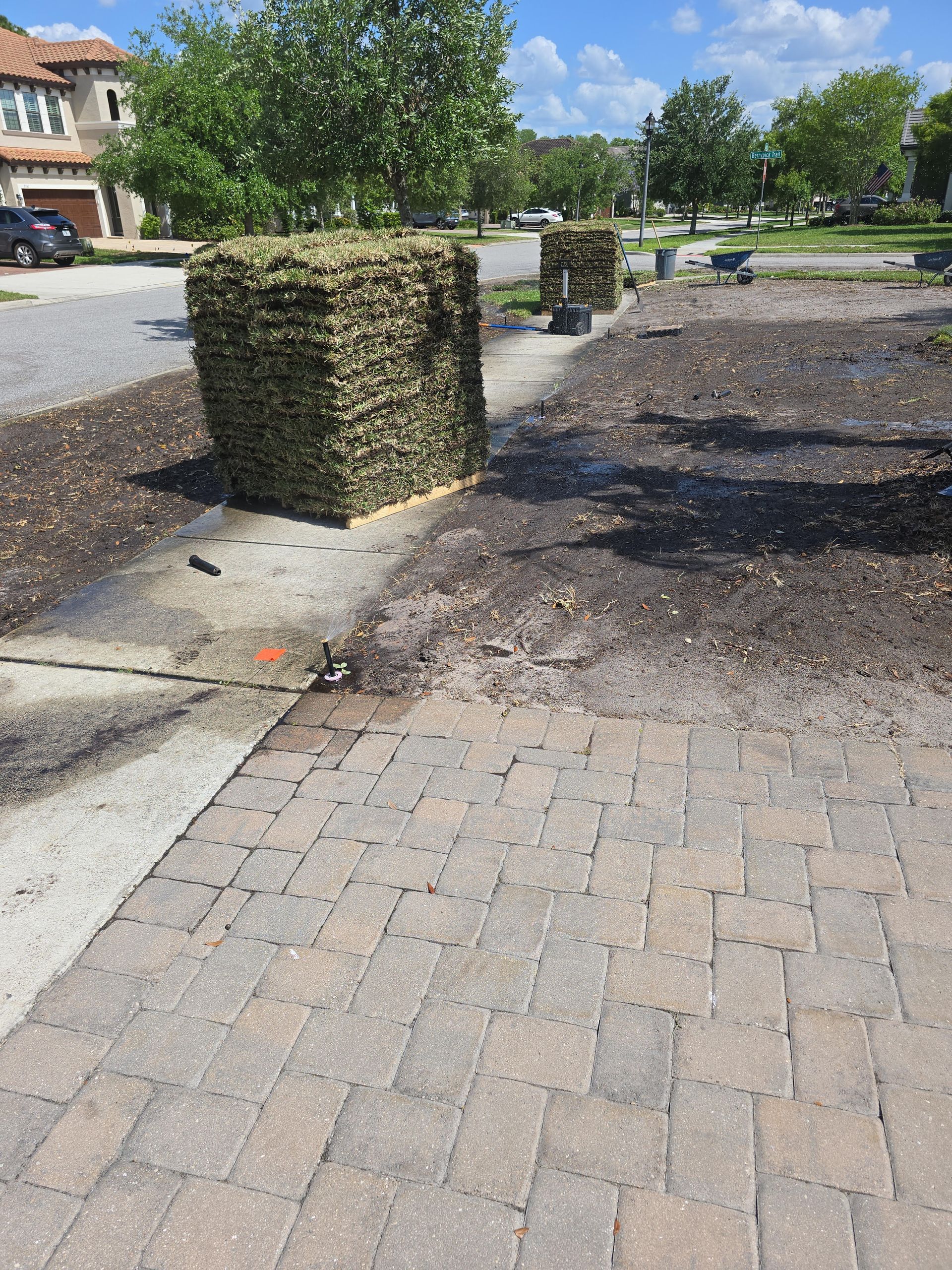 Two large, stacked hay bales sit on a paved driveway near a house, with a patch of dirt and mulch in the foreground.