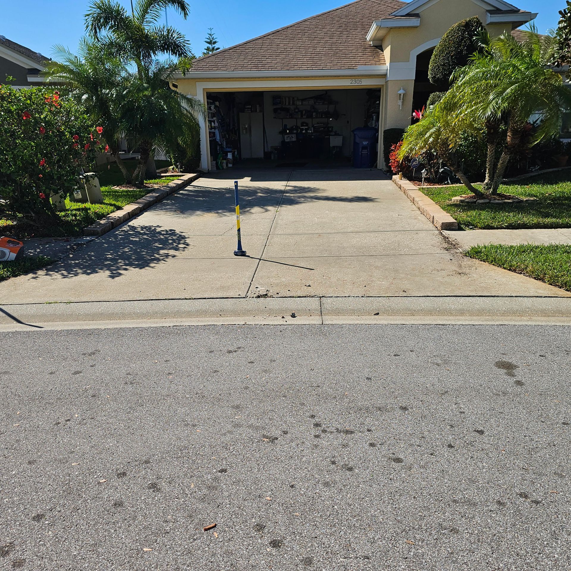 A concrete driveway leads to a suburban house garage with a blue and yellow tee ball stand centered in the path.