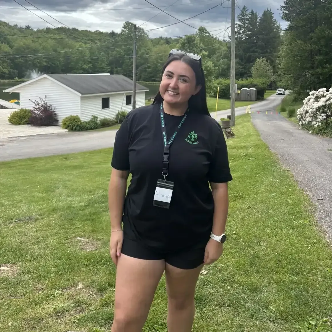 Woman with glasses and outstretched arms, smiling widely in a park. Red and white shirt with floral garland, sunny day.