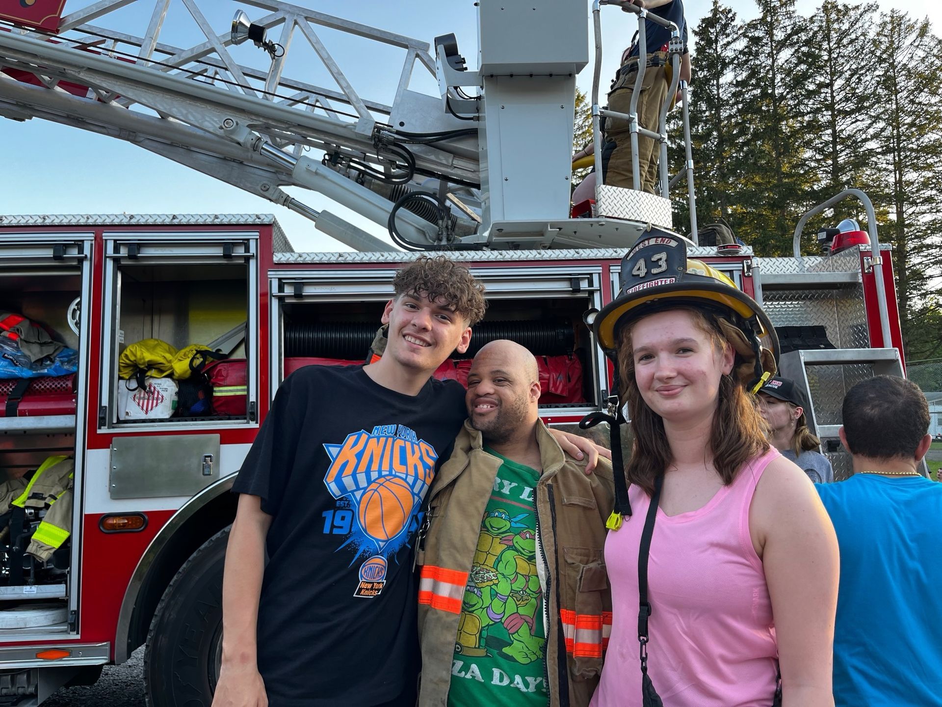 Three people smiling in front of a fire truck. The person in the center wears a firefighter coat.