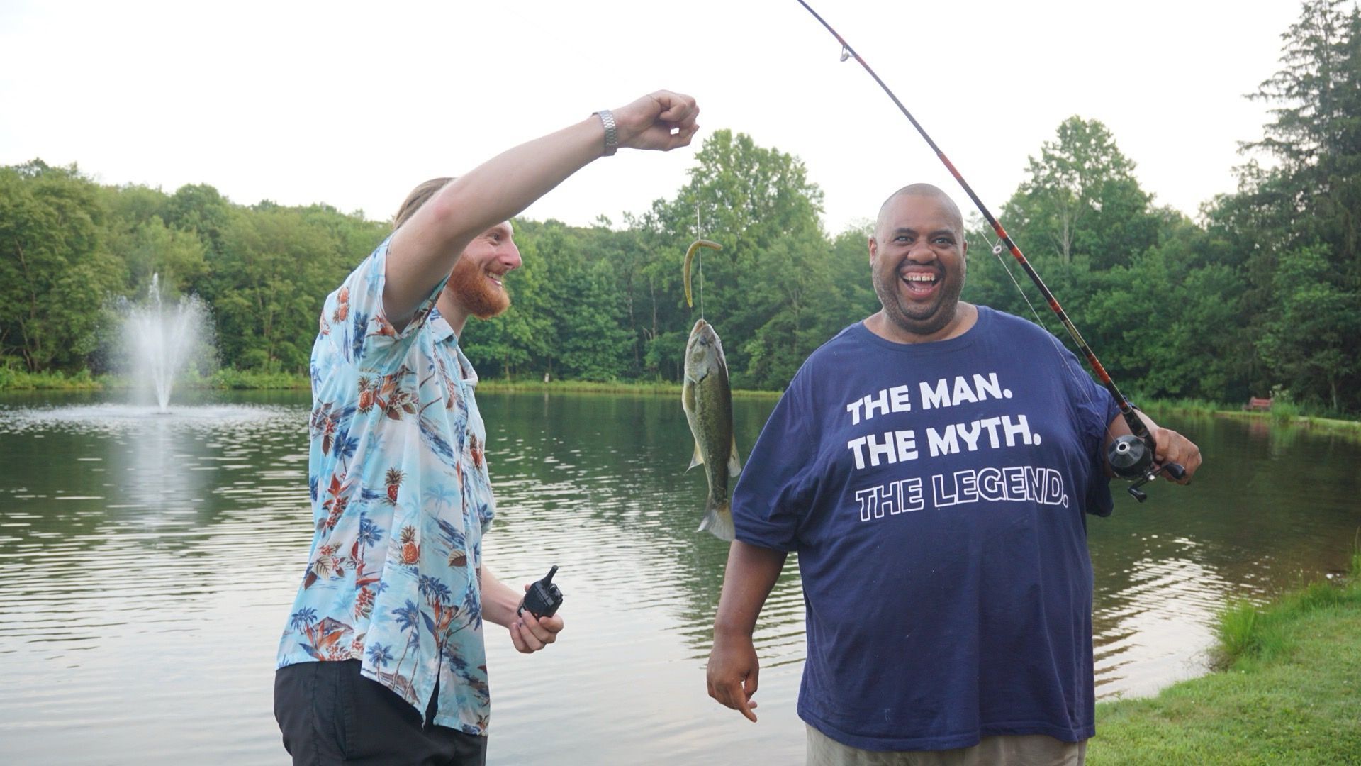 Two men fishing at a pond. One holds a pole with a fish, the other raises his arm in excitement.