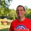 Man in red shirt with glasses smiles in park, bench in background.