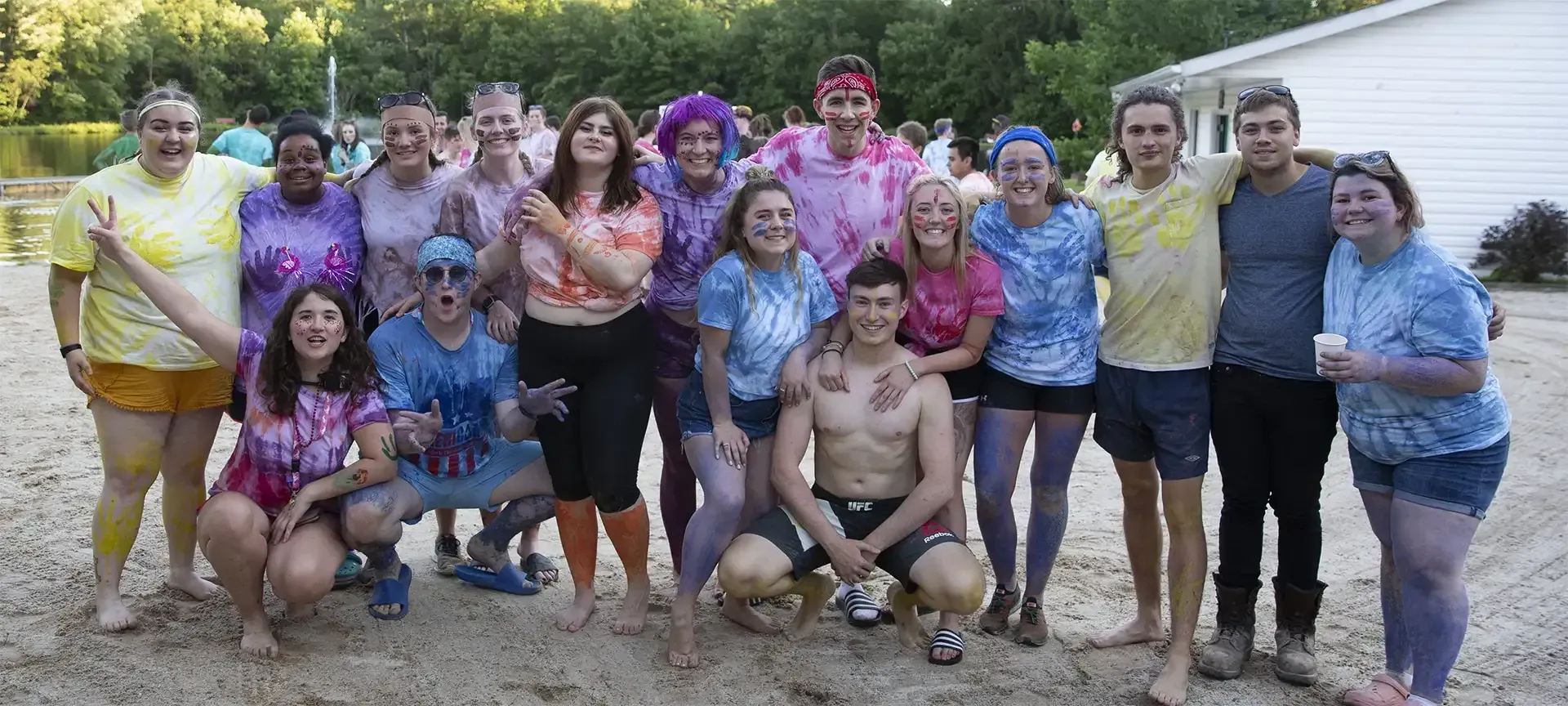 Group of people covered in colorful powder outdoors; tie-dye shirts, smiles, lake in background.