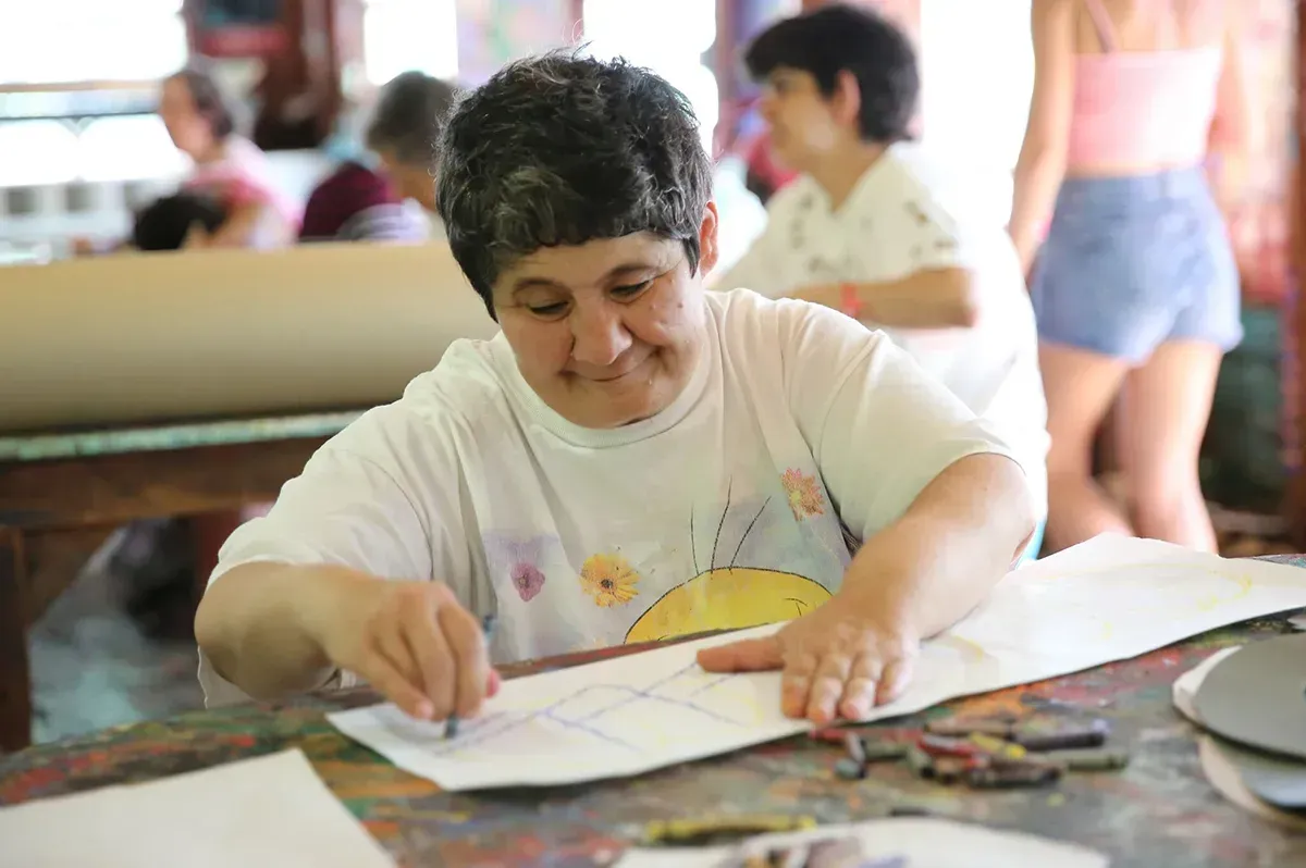 Woman drawing with crayons at a table, smiling. Other people visible in the background.