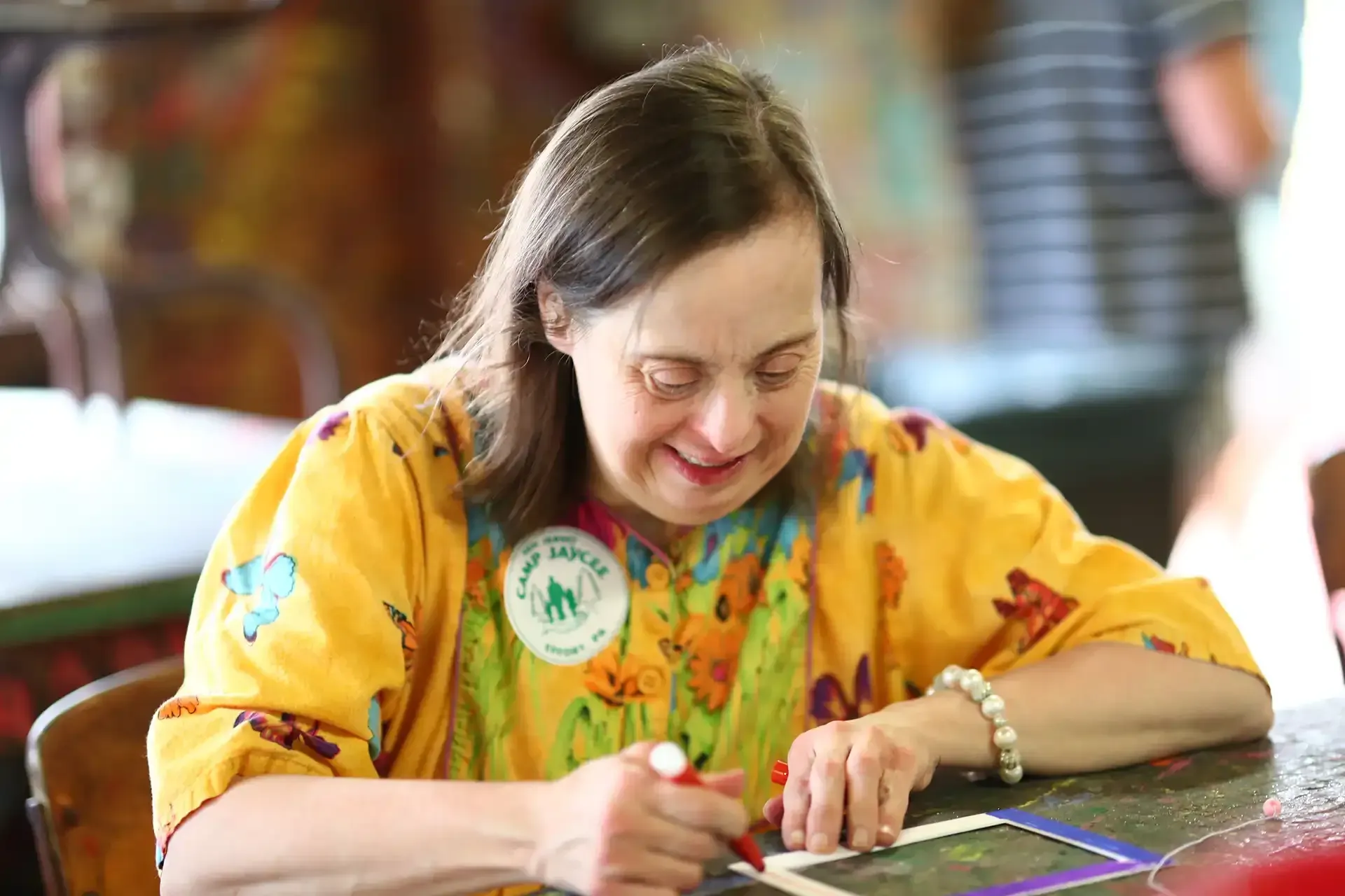 Woman in yellow shirt, smiling, writing on paper at a table. A button on her shirt.