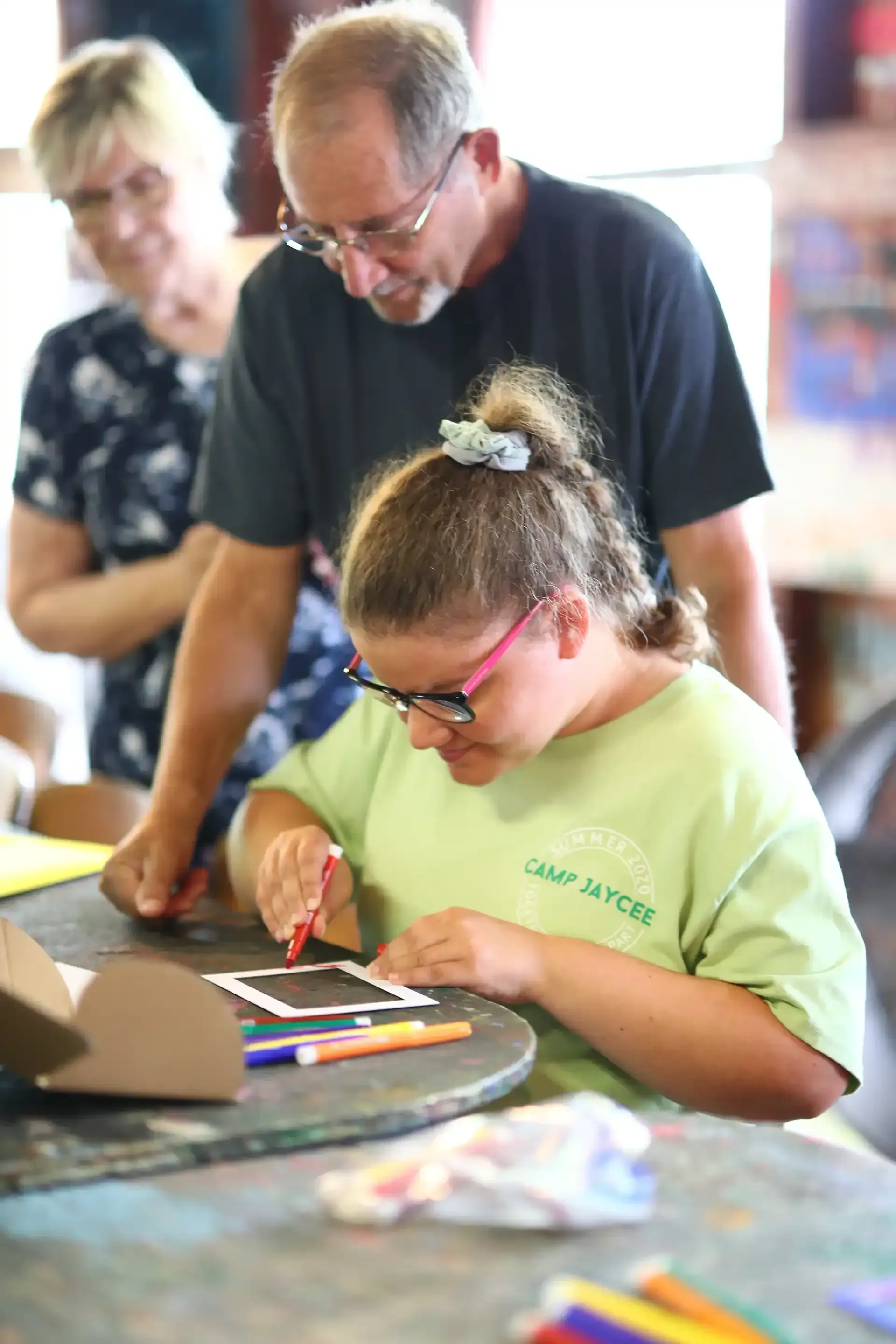 A person with glasses drawing at a table with help from an instructor, others watch.