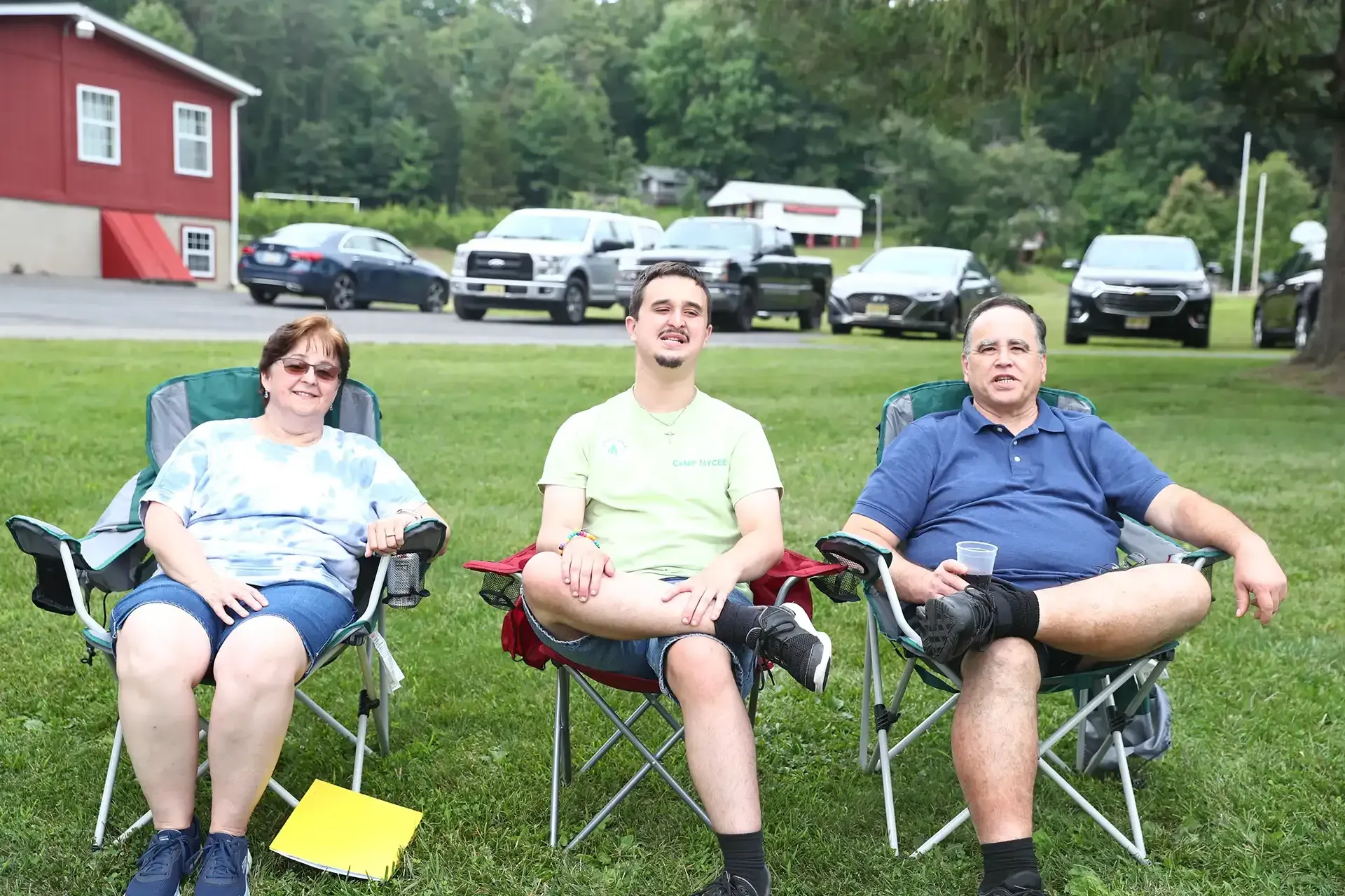 Three people sitting in folding chairs on a grassy lawn with cars and a building in the background.
