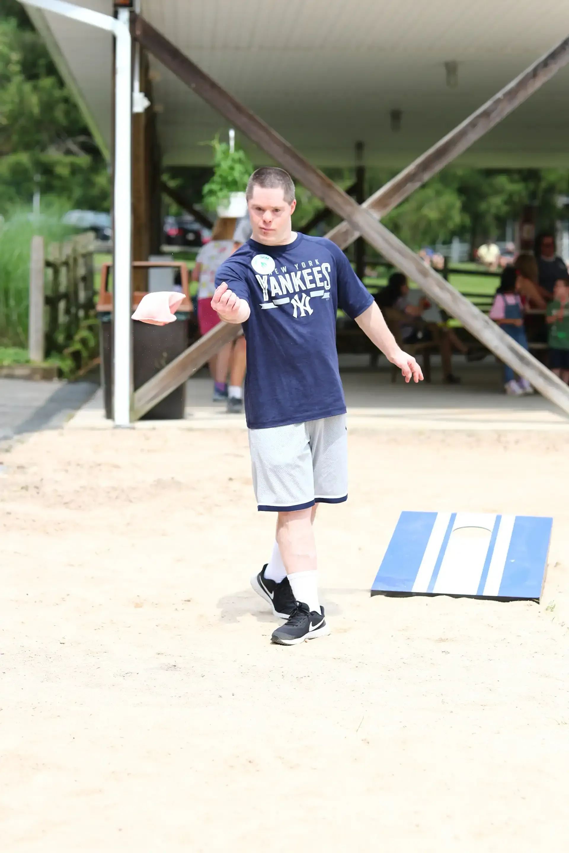 Man in Yankees shirt throws beanbag at a cornhole board in an outdoor setting.