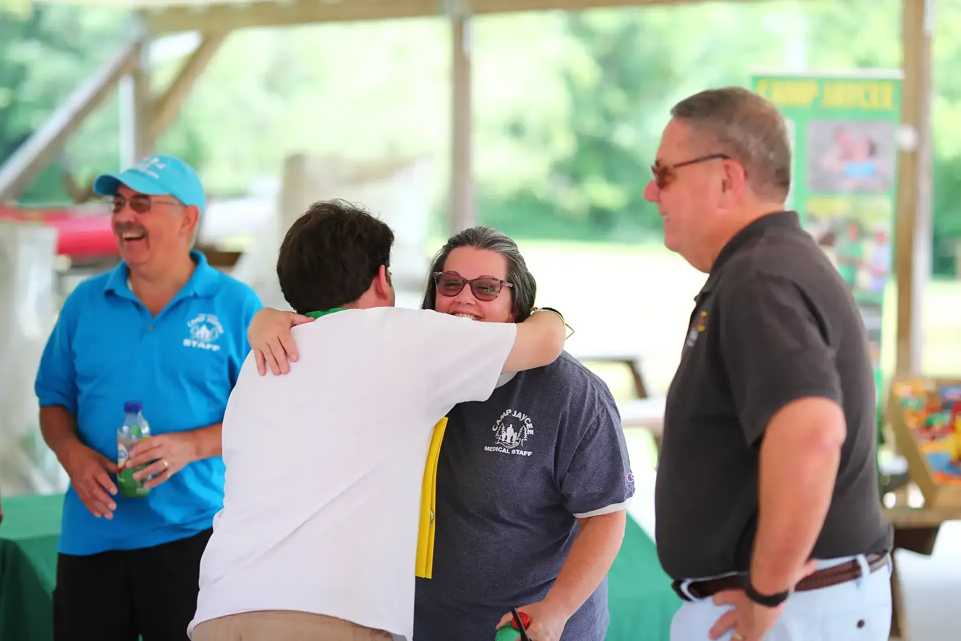 Two people hugging under a shelter; two others look on smiling. Green table, outdoor setting.