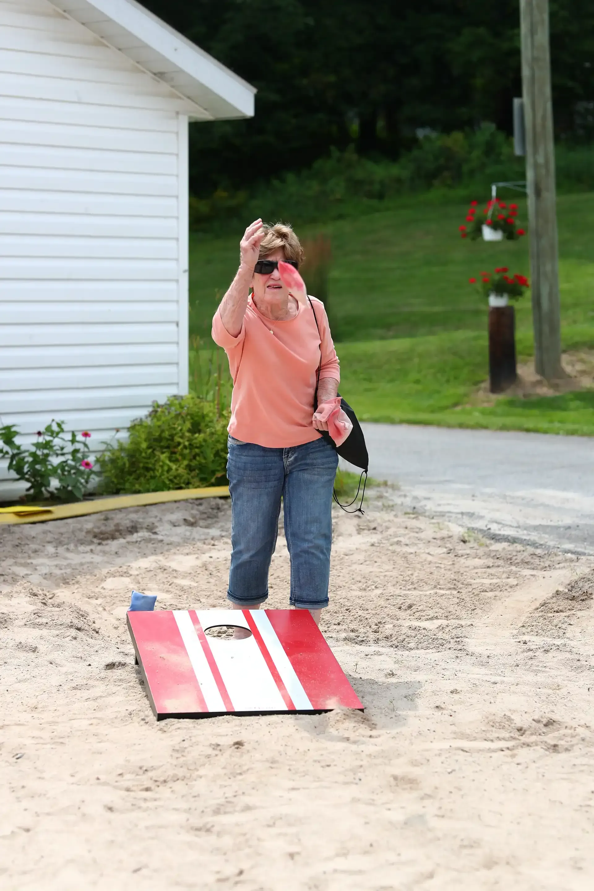 Woman tossing a bean bag toward a cornhole board on a sandy area.