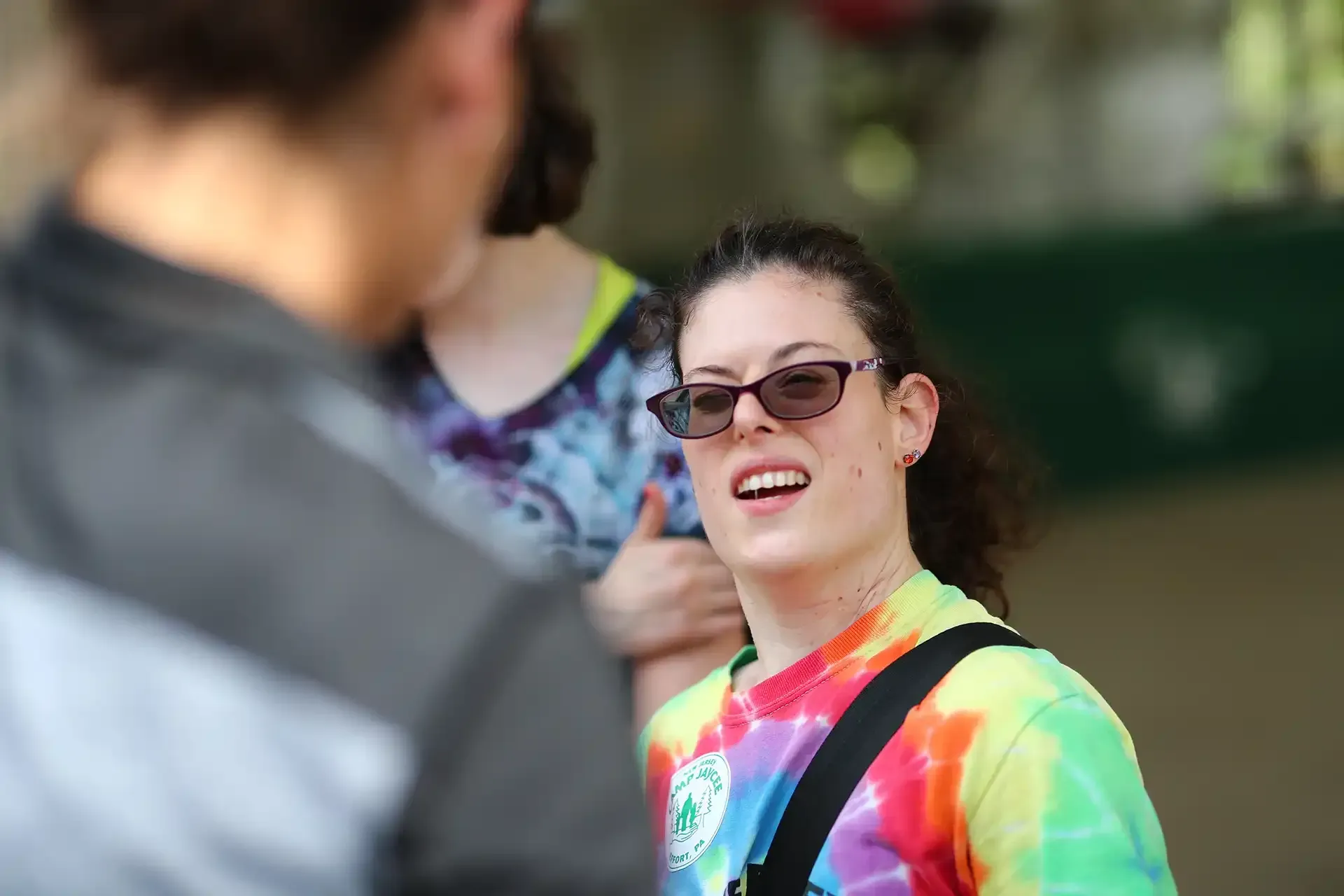 Woman with sunglasses and tie-dye shirt talking outdoors, with another person in the foreground and another behind.