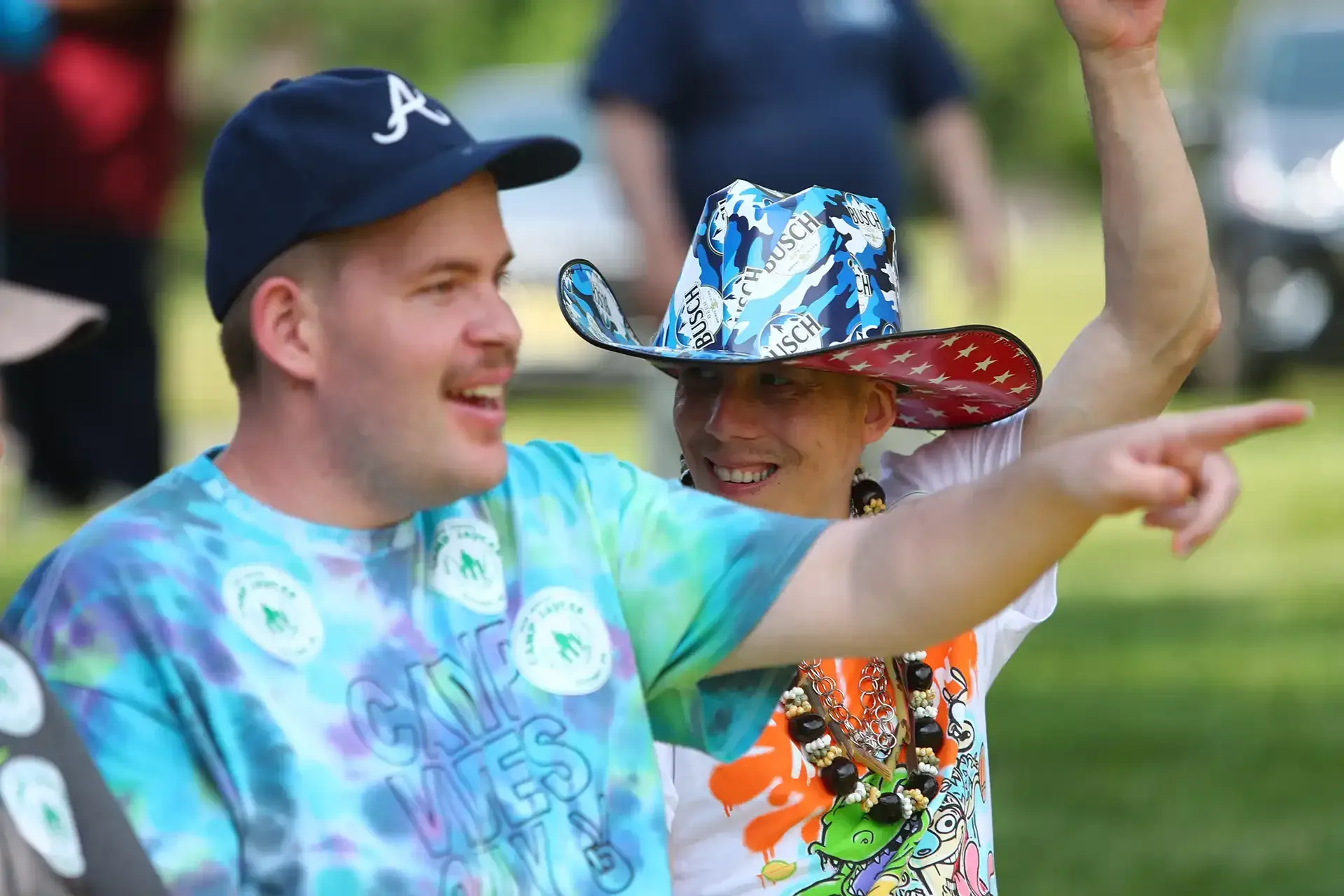 Two people smiling and pointing outdoors. One wears a tie-dye shirt and a baseball cap; the other, a cowboy hat.