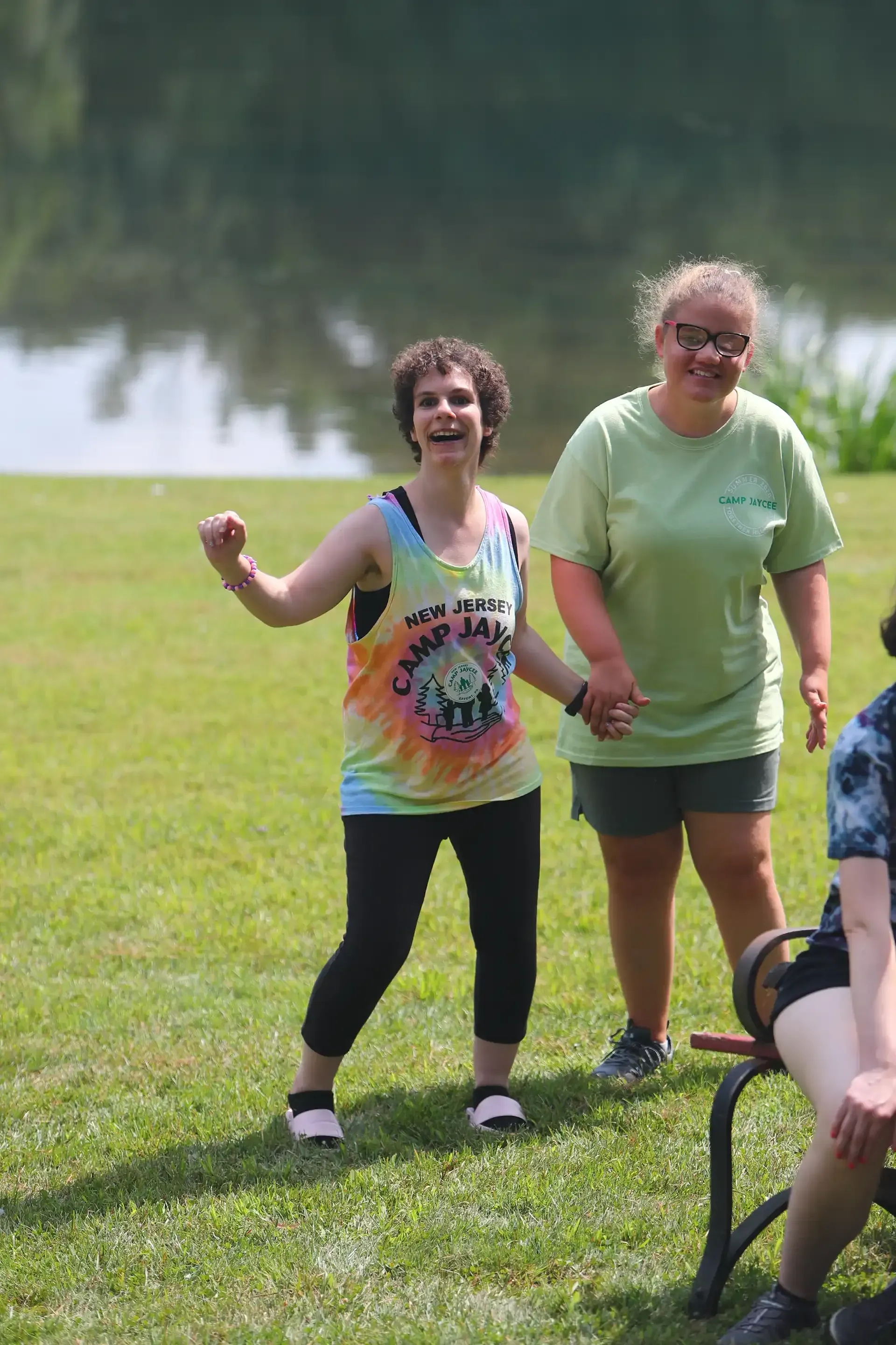 Two people smiling and holding hands on a grassy field near water. One wears a tie-dye shirt.