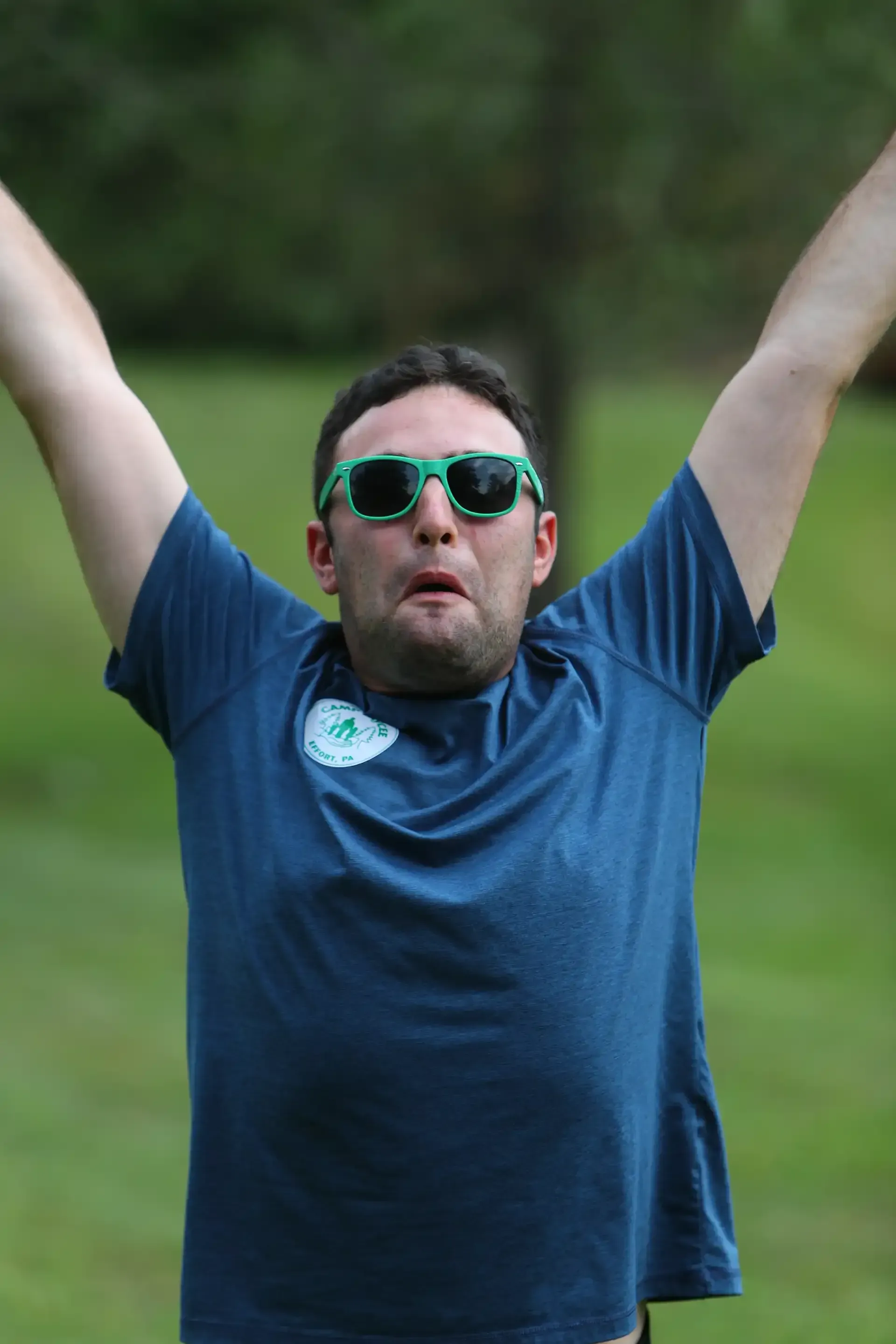 Man wearing green sunglasses with arms raised and mouth open outdoors.