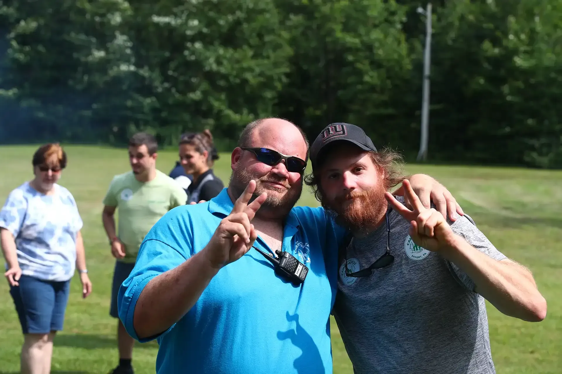 Two men smile, flashing peace signs, embraced in a green field. Others stand in background.
