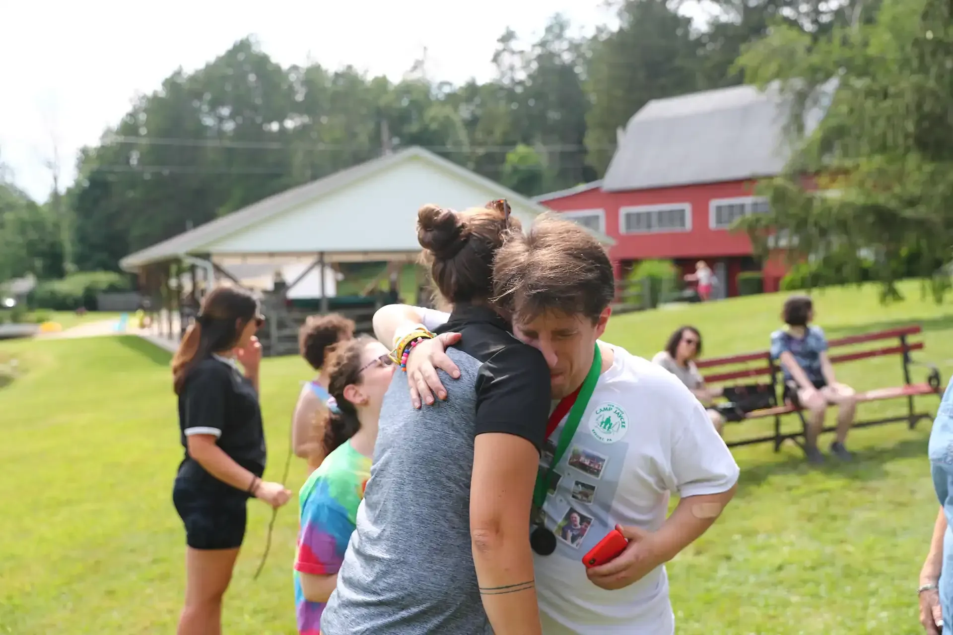 People hugging outdoors; person wearing medal and hugging. Green grass, a barn, and trees in the background.