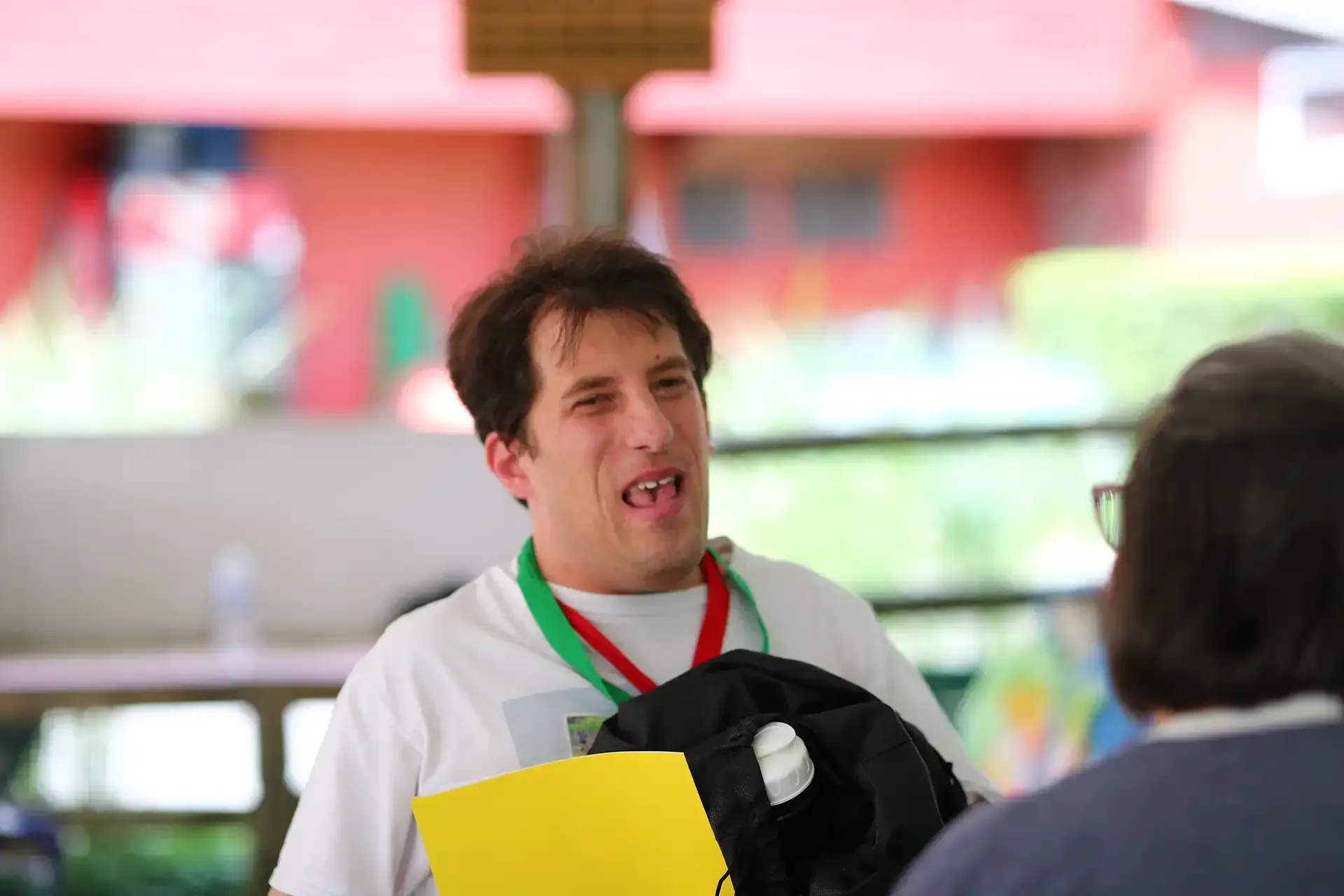 Man with lanyard smiling, holding yellow paper and black bag, speaking to person off-camera, outdoor setting.