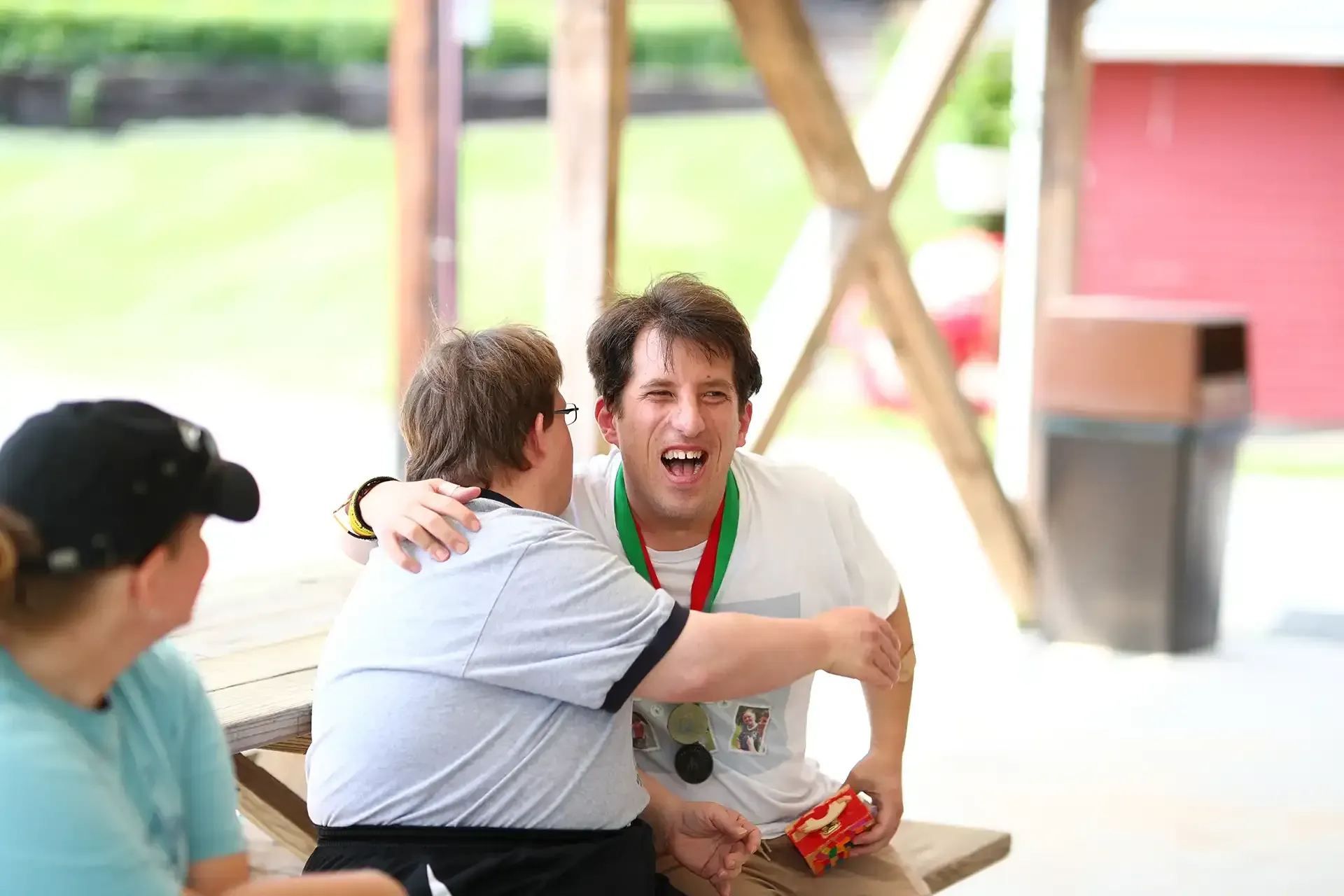 Two people embrace outdoors; one wearing a medal, smiling widely. A third person sits nearby.