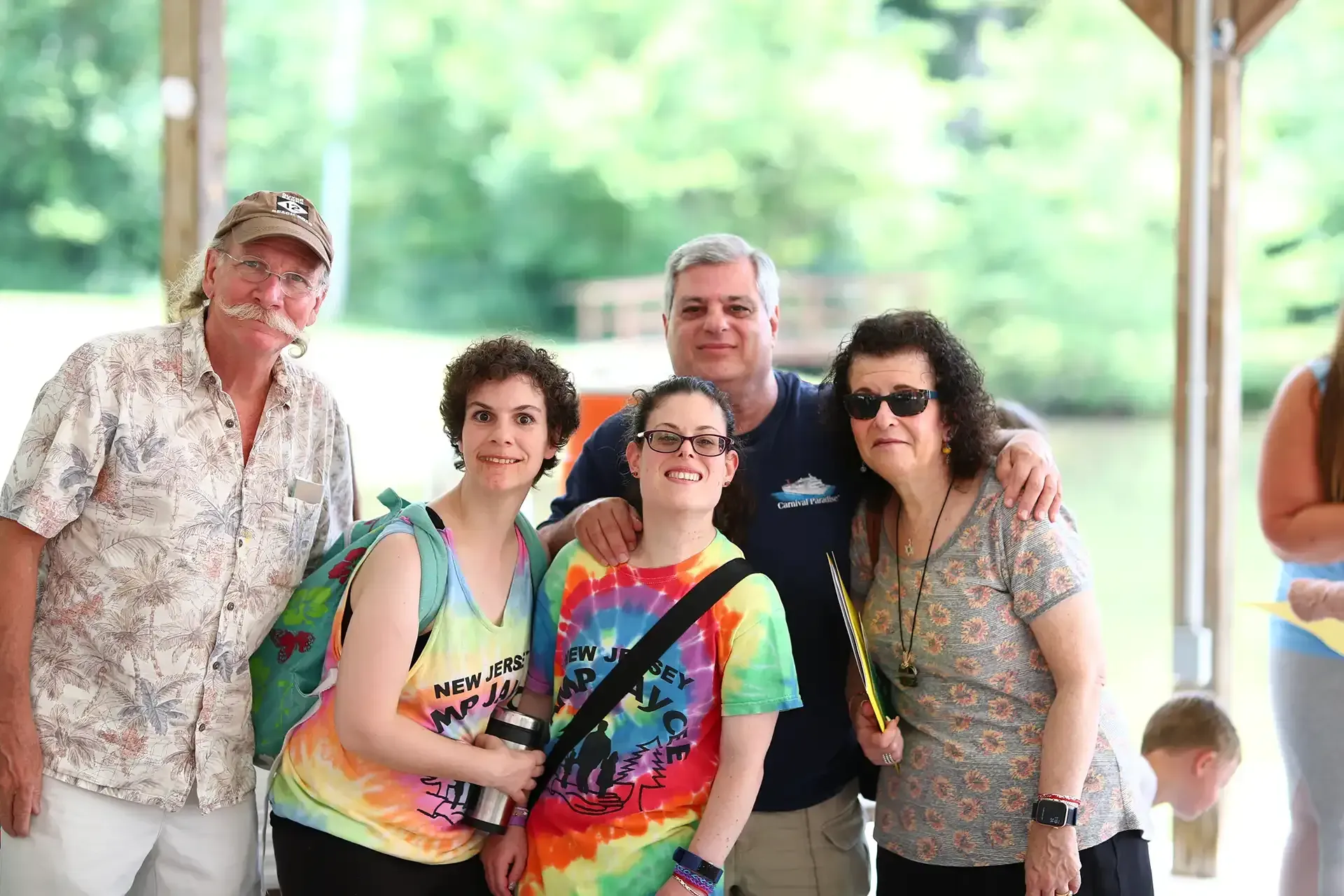 Group of five people smiling outdoors by water; one wears tie-dye, another sunglasses.