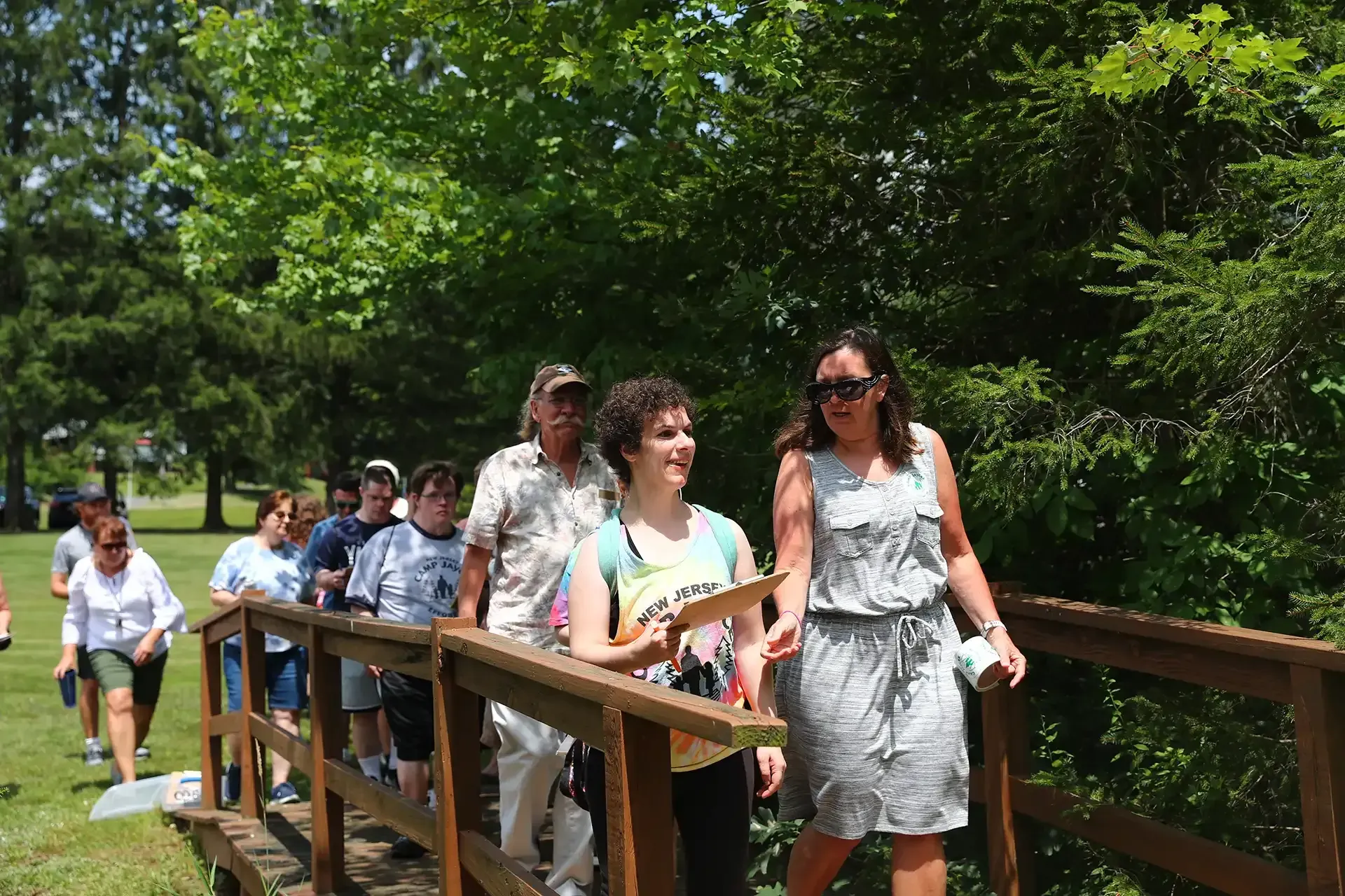 Group of people walking on a wooden bridge outdoors, sunny day. Trees in the background.