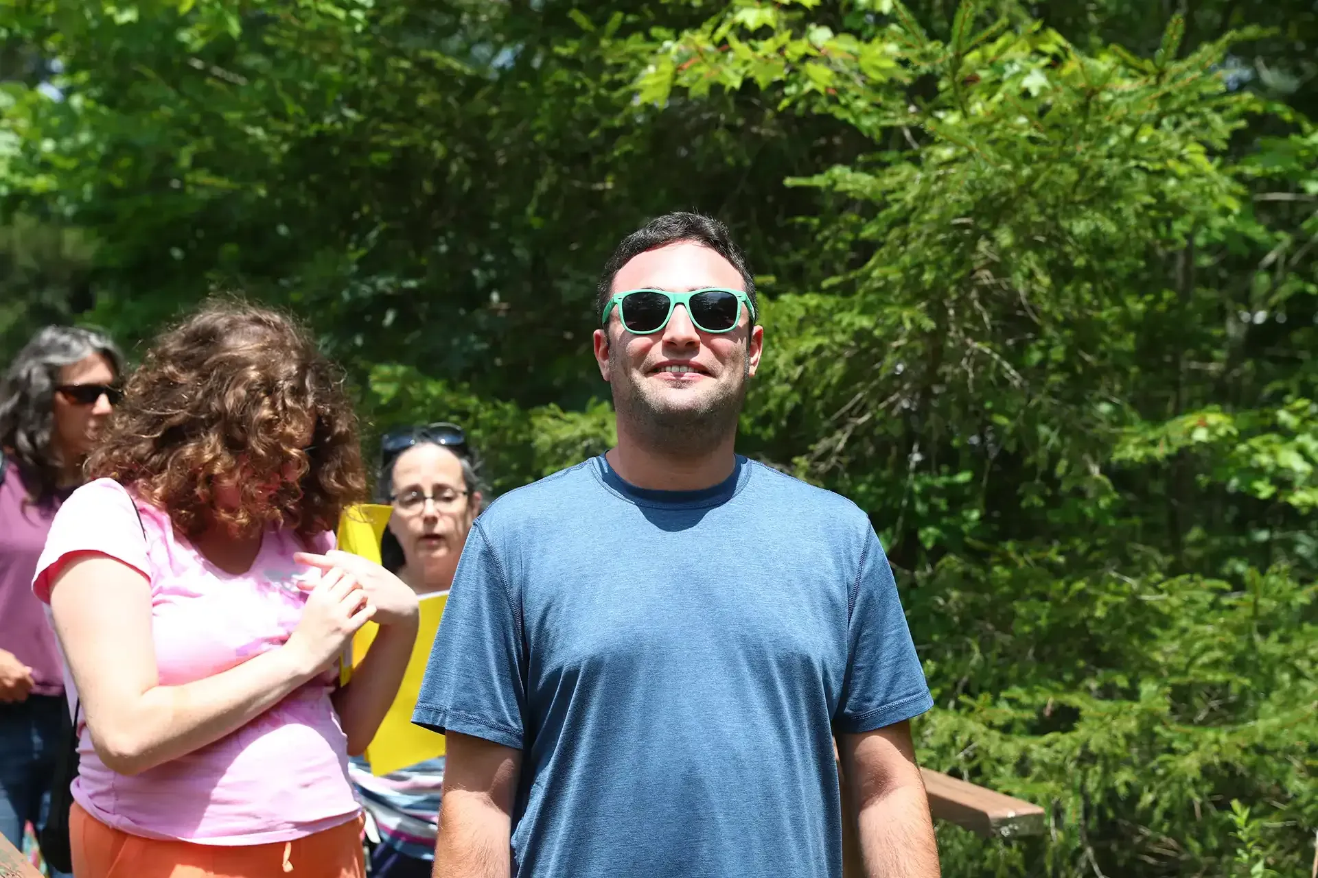 Man in blue shirt and sunglasses smiles, walking outdoors with a group of people.