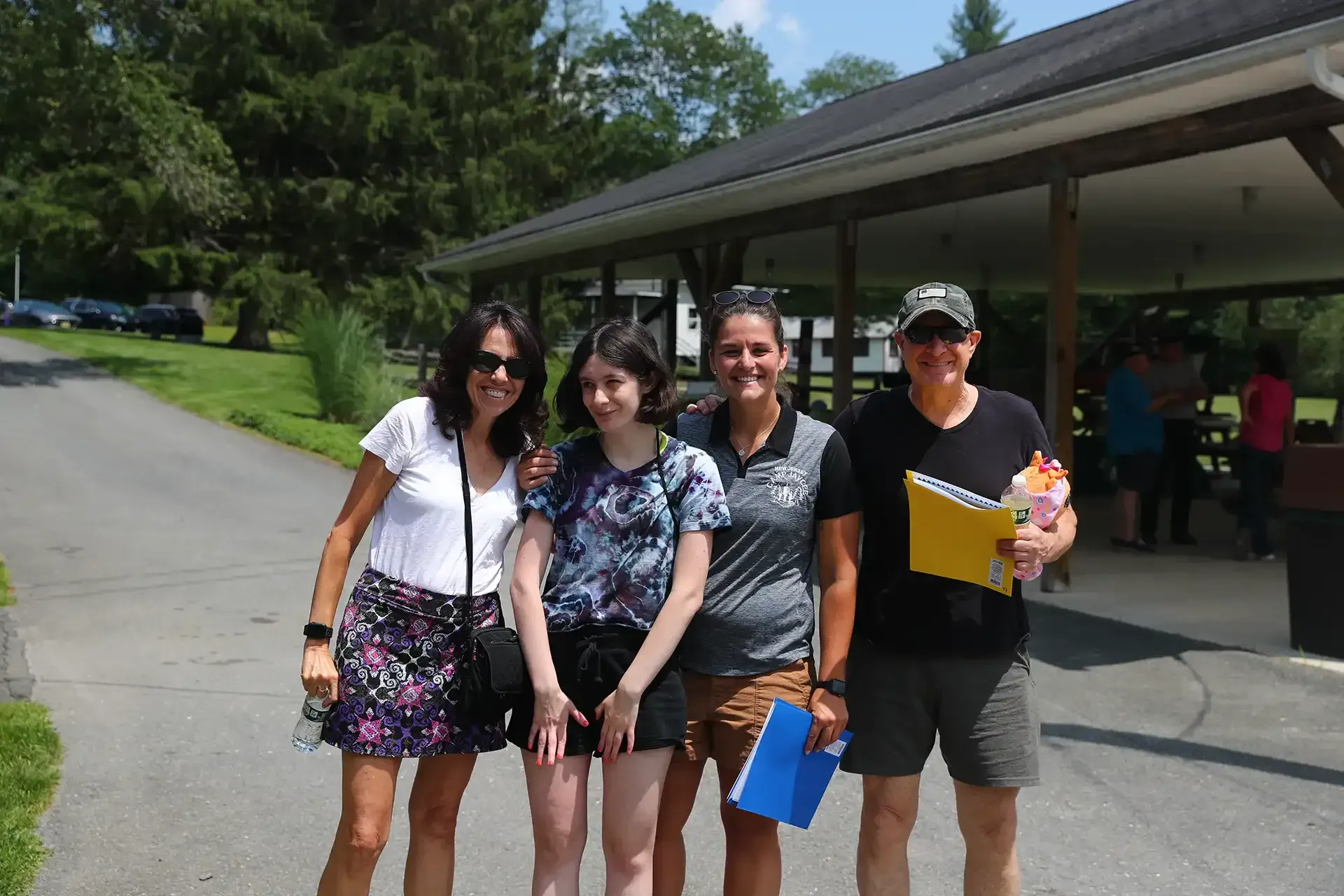 Four people posing outdoors under a pavilion. One holds a yellow notebook and small gift. Others smile.