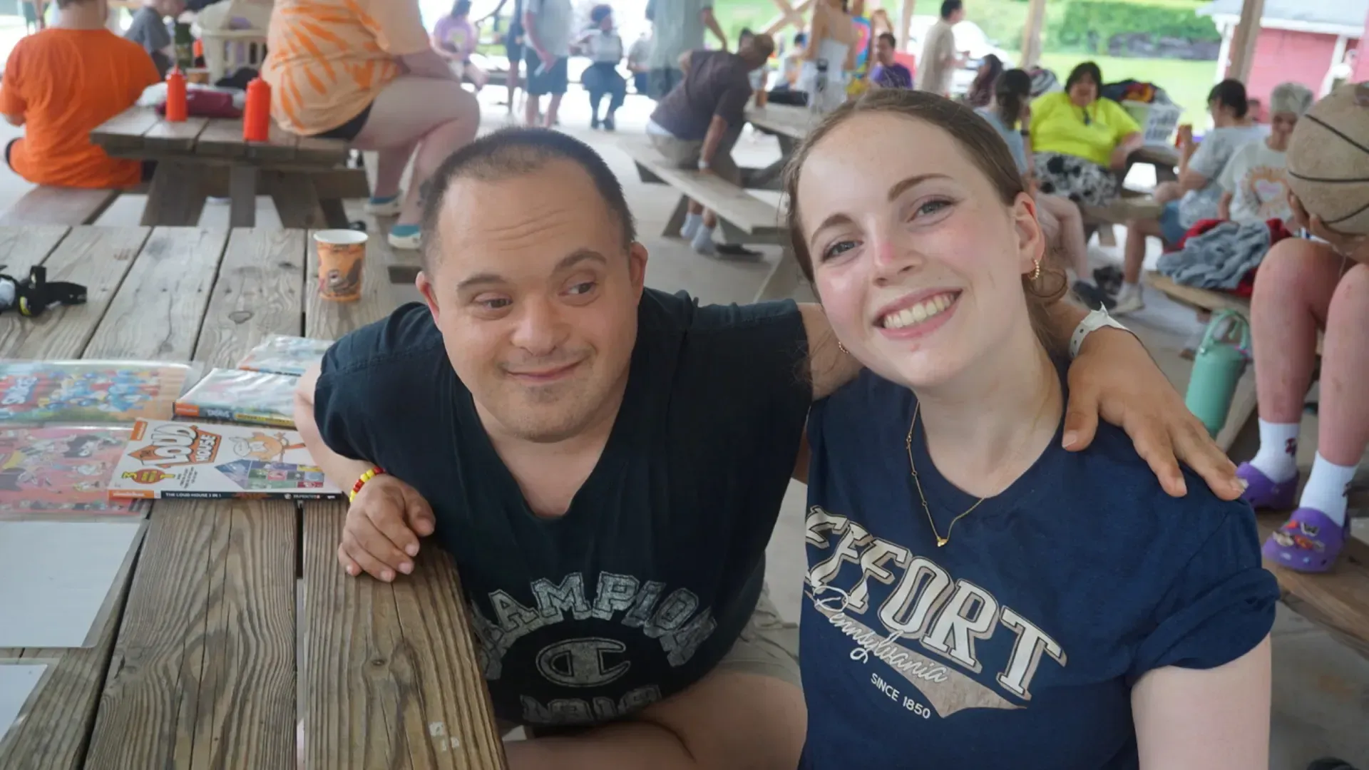Two people smiling at picnic table. Man with Down syndrome arm around woman. Others seated nearby.