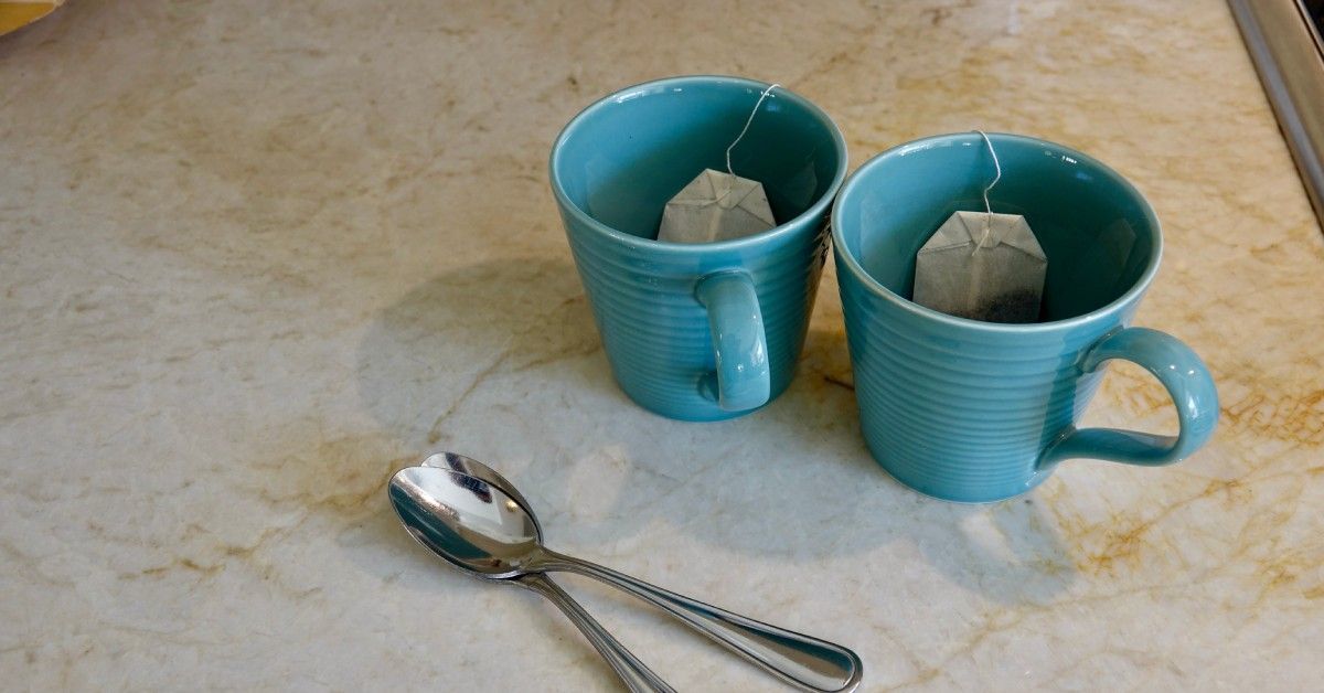 Two aquamarine teacups with teabags and two silver spoons rest on a lovely white quartzite countertop.