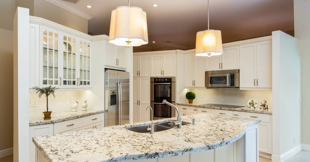 A lovely white kitchen with overhead lamps and a grey and white granite tabletop with a sink in the middle.