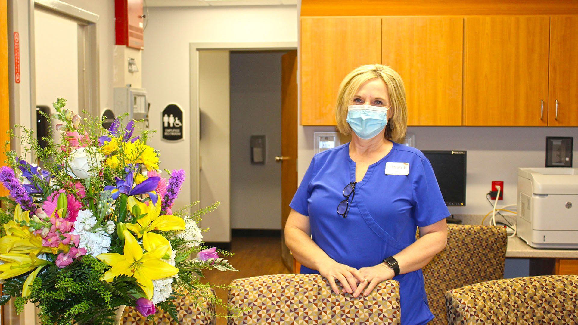 Woman in blue scrubs and mask stands by a reception desk, next to a large flower arrangement.