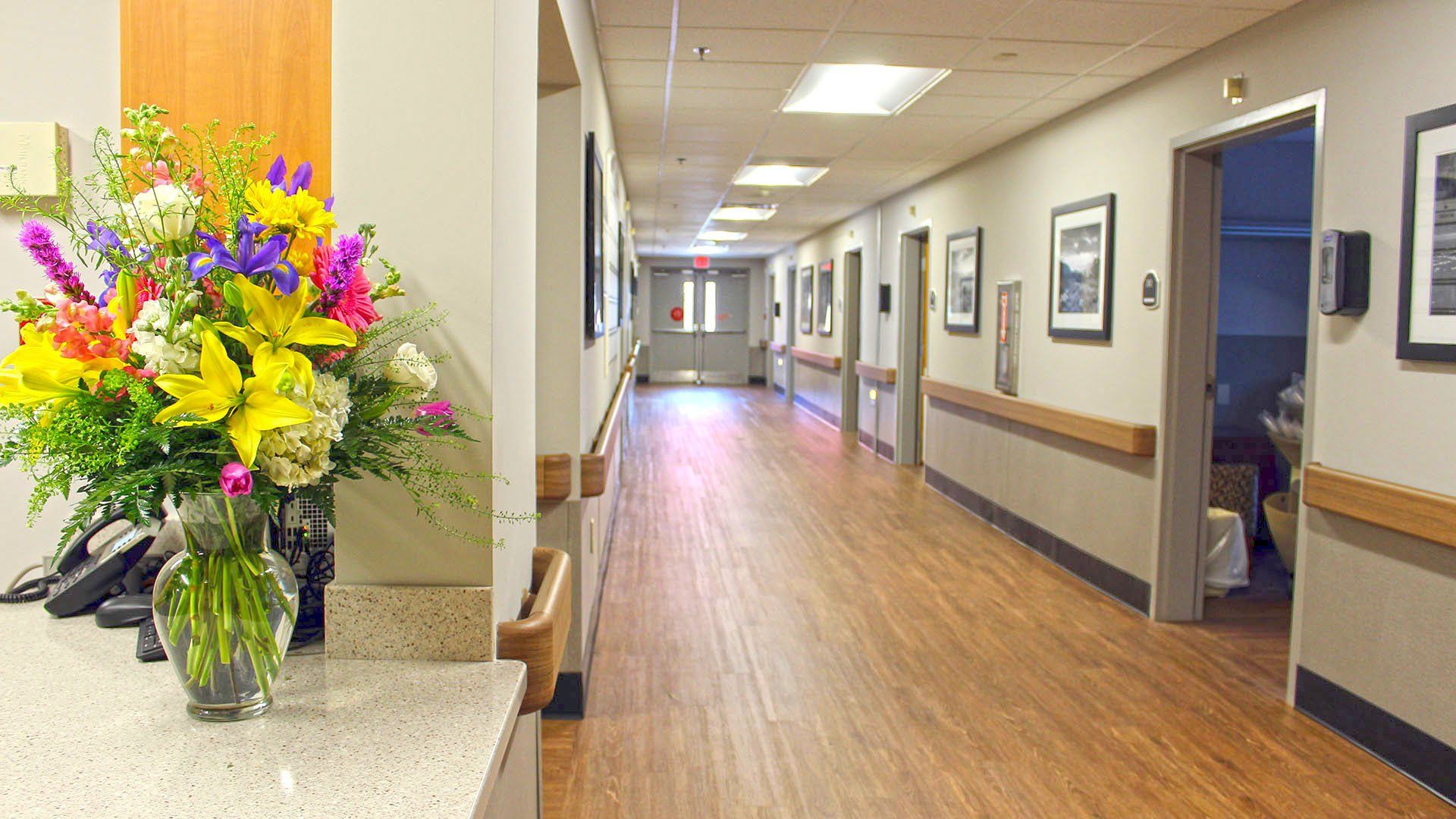 Long hallway in a building with wooden floors, neutral-colored walls, and framed pictures. Flowers sit on a counter on the left.