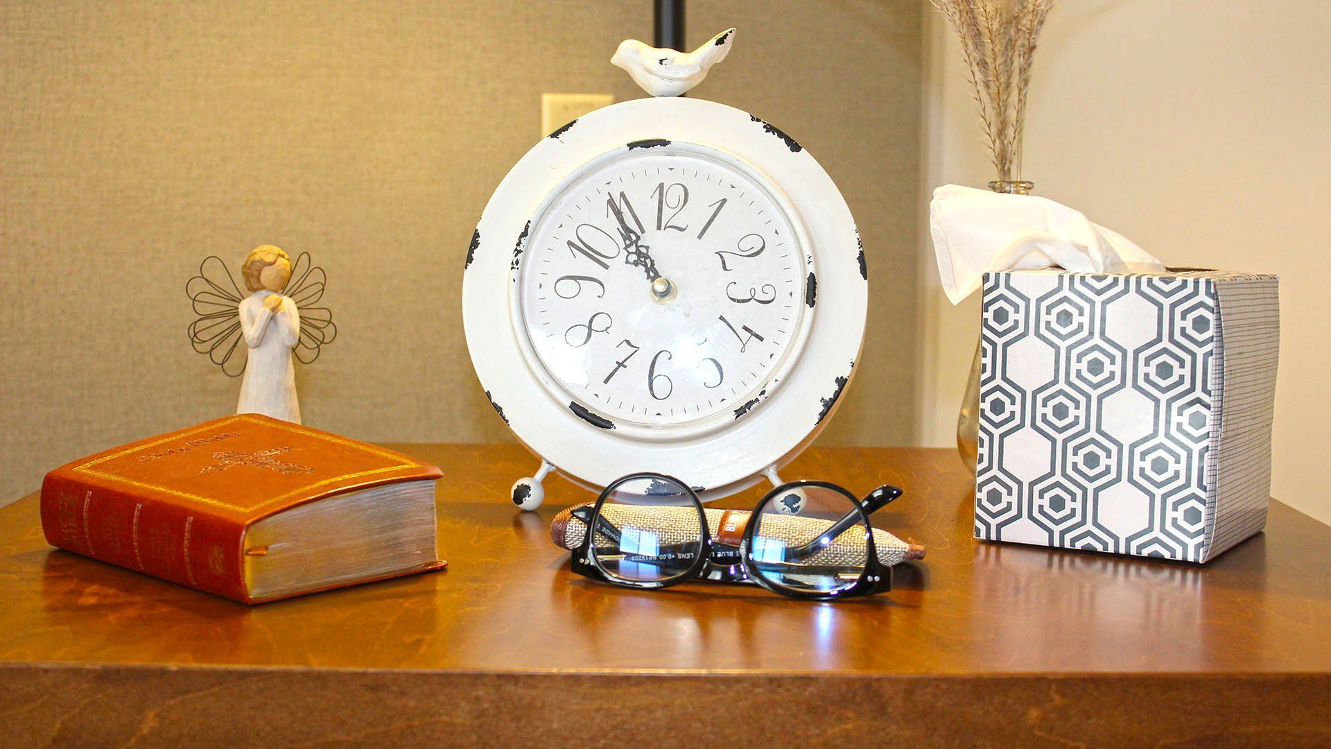 White clock, glasses, book, angel figurine, and tissue box on a brown wooden surface.